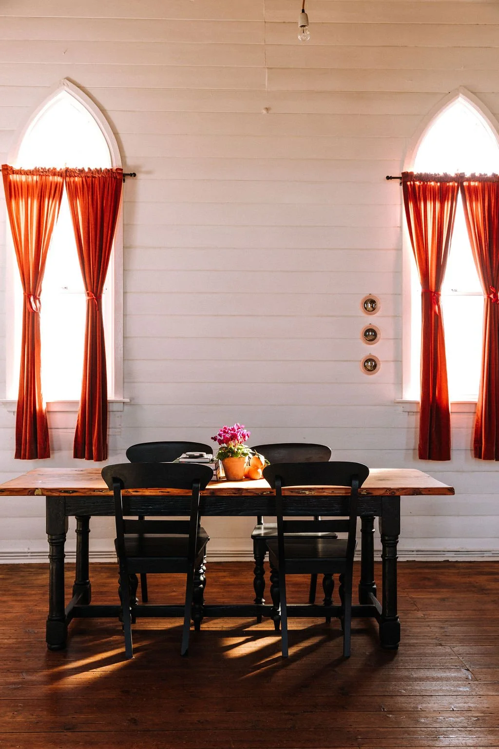 A dining area with a wooden table and four black chairs, a flowerpot with pink flowers, and some apples. The background features white wooden paneled walls, three circular wall decorations, and two tall arched windows with red curtains, allowing natu