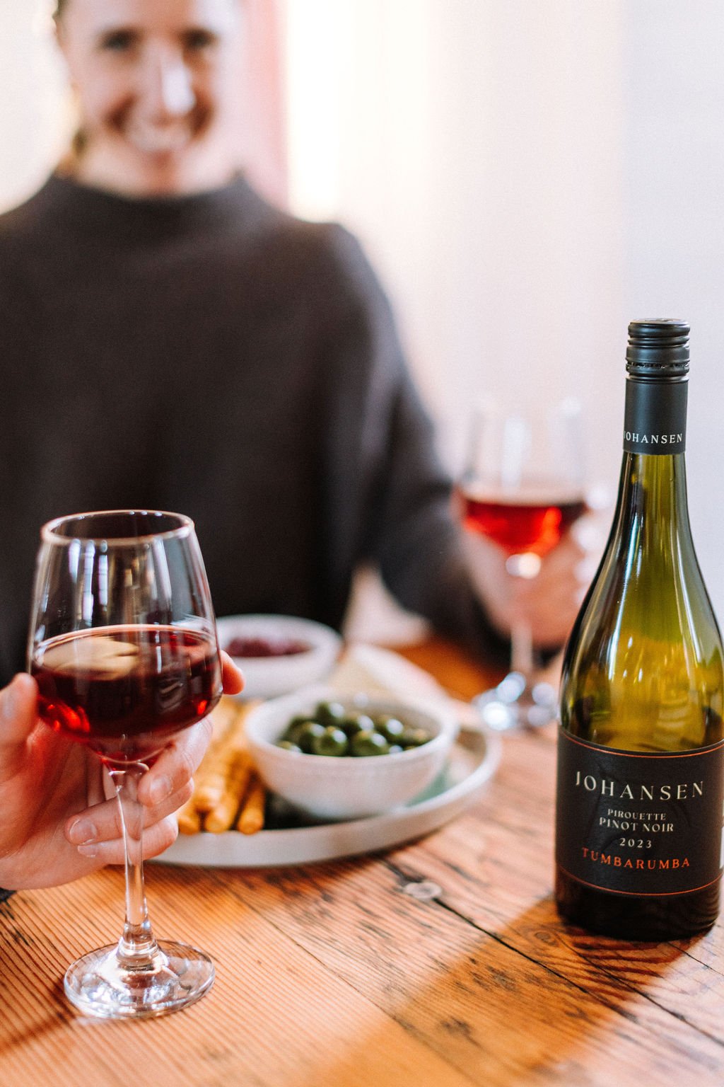 People enjoying a wine tasting with glasses of red wine on a wooden table, accompanied by bowls of snacks and a bottle of Johansen Tumbarrumba Pinot Noir 2023.
