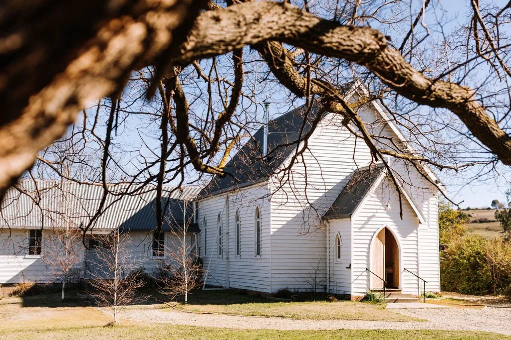 A white wooden church with a steeple, framed by tree branches in the foreground, set in a rural area with a grassy yard and distant hills.