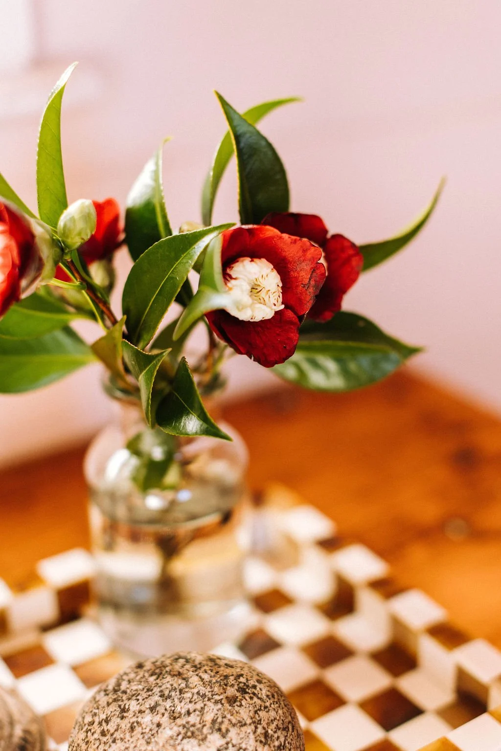 A small bouquet of flowers with red and white blooms and green leaves in a glass vase on a wooden surface with a checkered mat and rounded stones nearby.