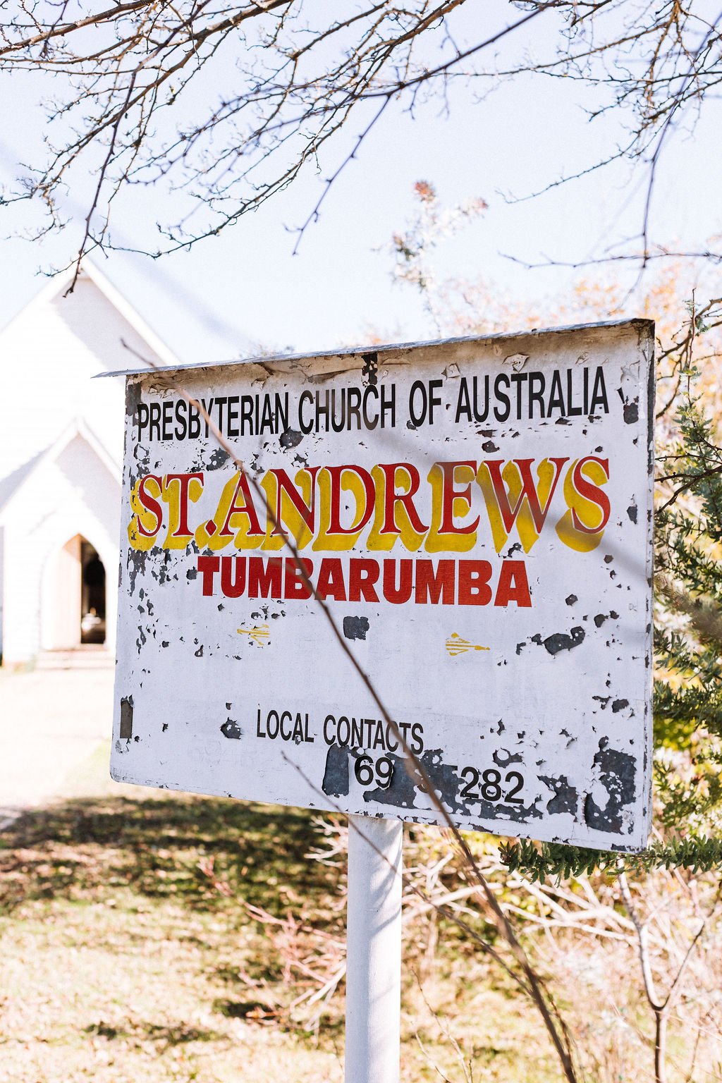 An aged sign for the Presbyterian Church of Australia and St. Andrews Tumbarumba, with a church building partially visible in the background, and leafless tree branches above.