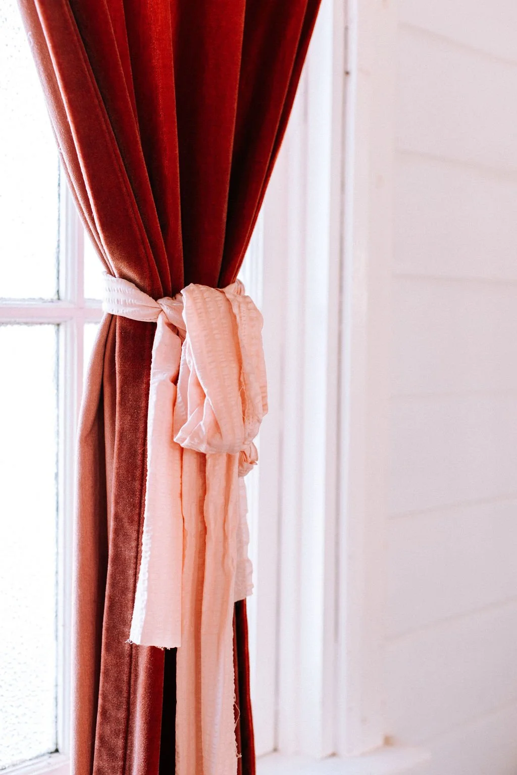Close-up of a red velvet curtain tied back with a pink ribbon, next to a window with frosted glass, and a white wall.