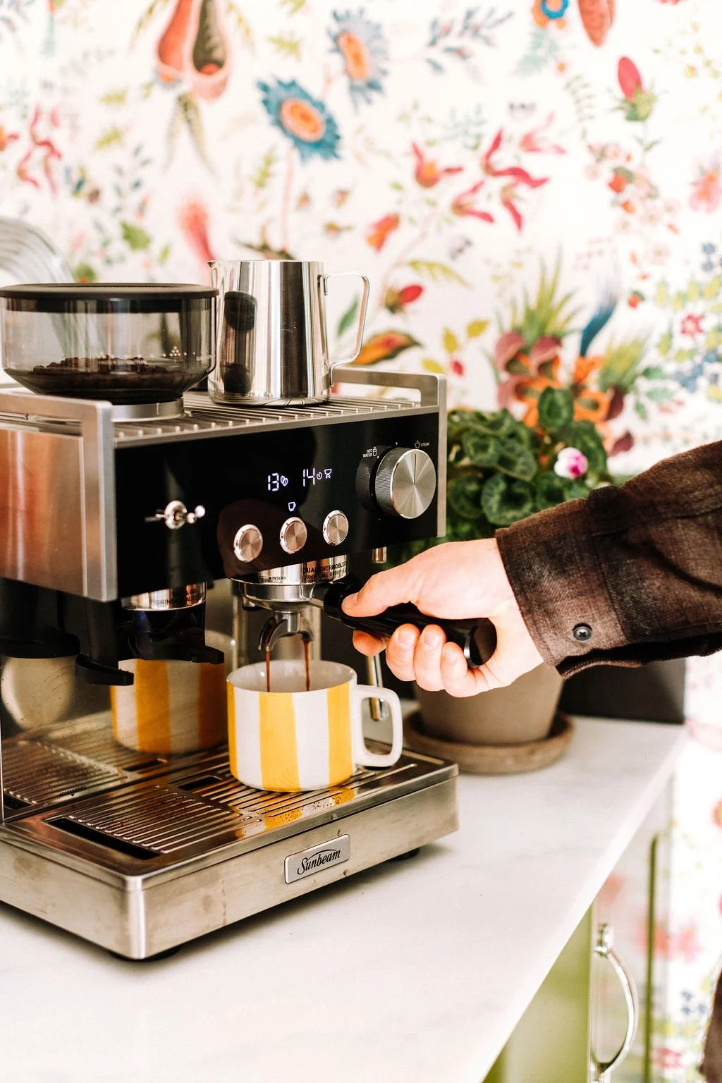 Person making espresso with a coffee machine and yellow-striped mug in a cozy, floral-themed kitchen.