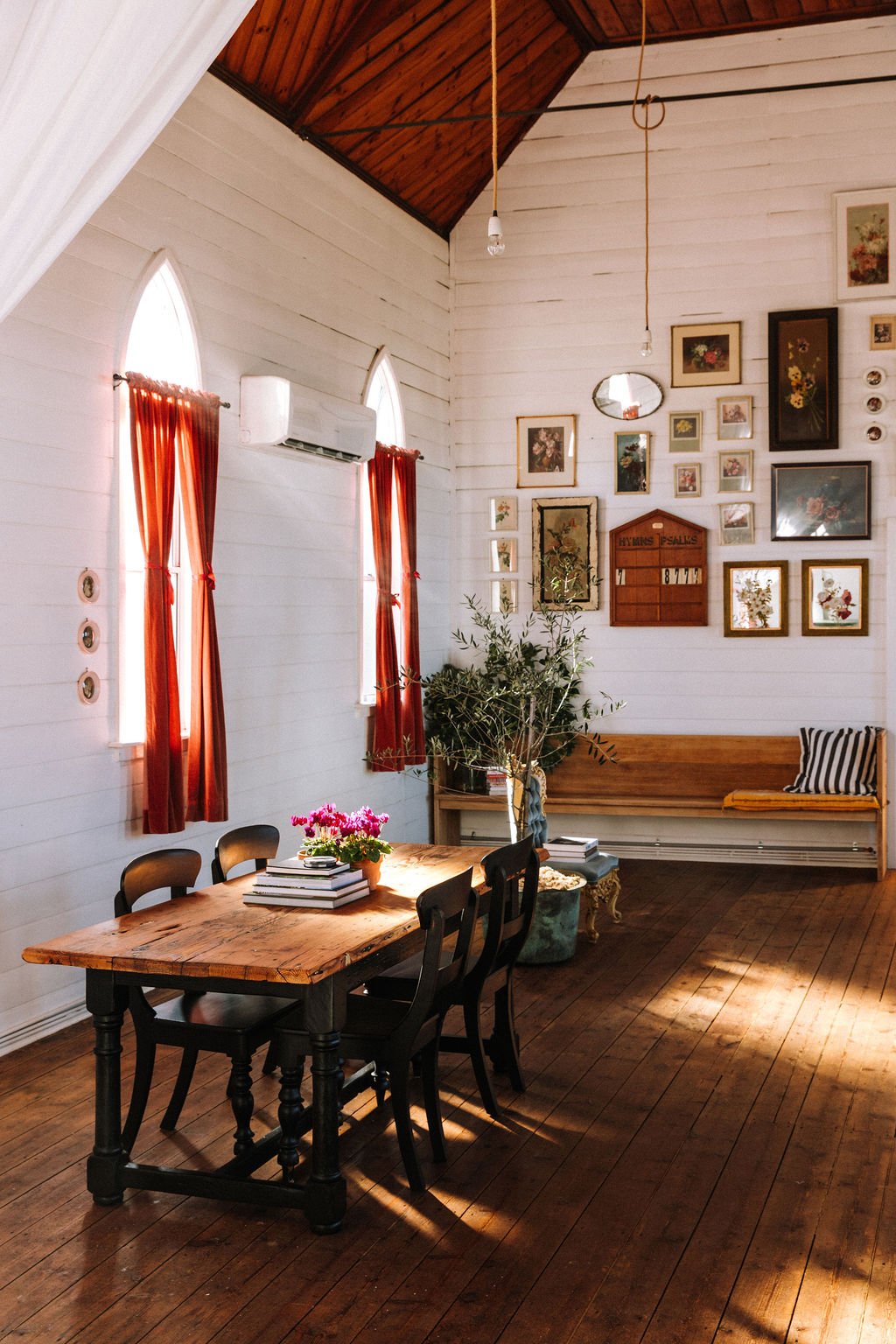 Sunlit dining area with a wooden table, black chairs, and a vase of pink flowers. White wooden walls decorated with framed artwork and a wall-mounted mirror. Red curtains on arched windows, and a wooden bench with striped pillow. Indoor plants add gr