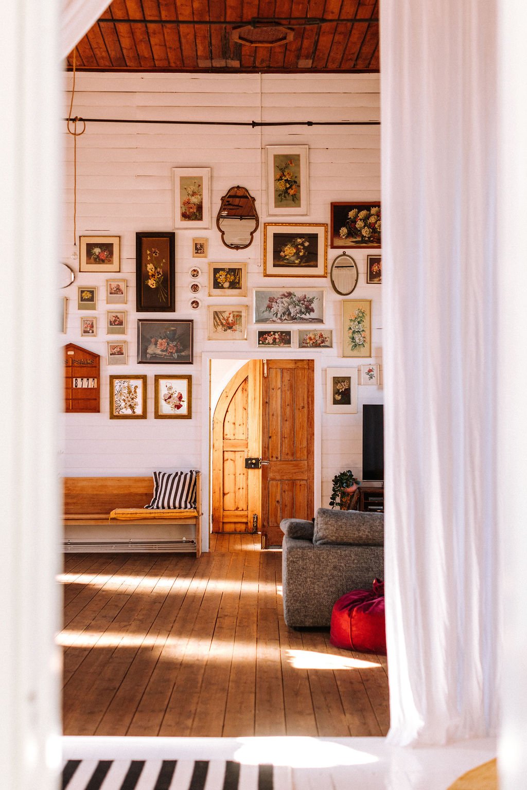 View of a cozy living room with a gallery wall of framed floral art, a wooden bench with striped cushion, a gray sofa, and a partially open wooden door, seen through white curtains.