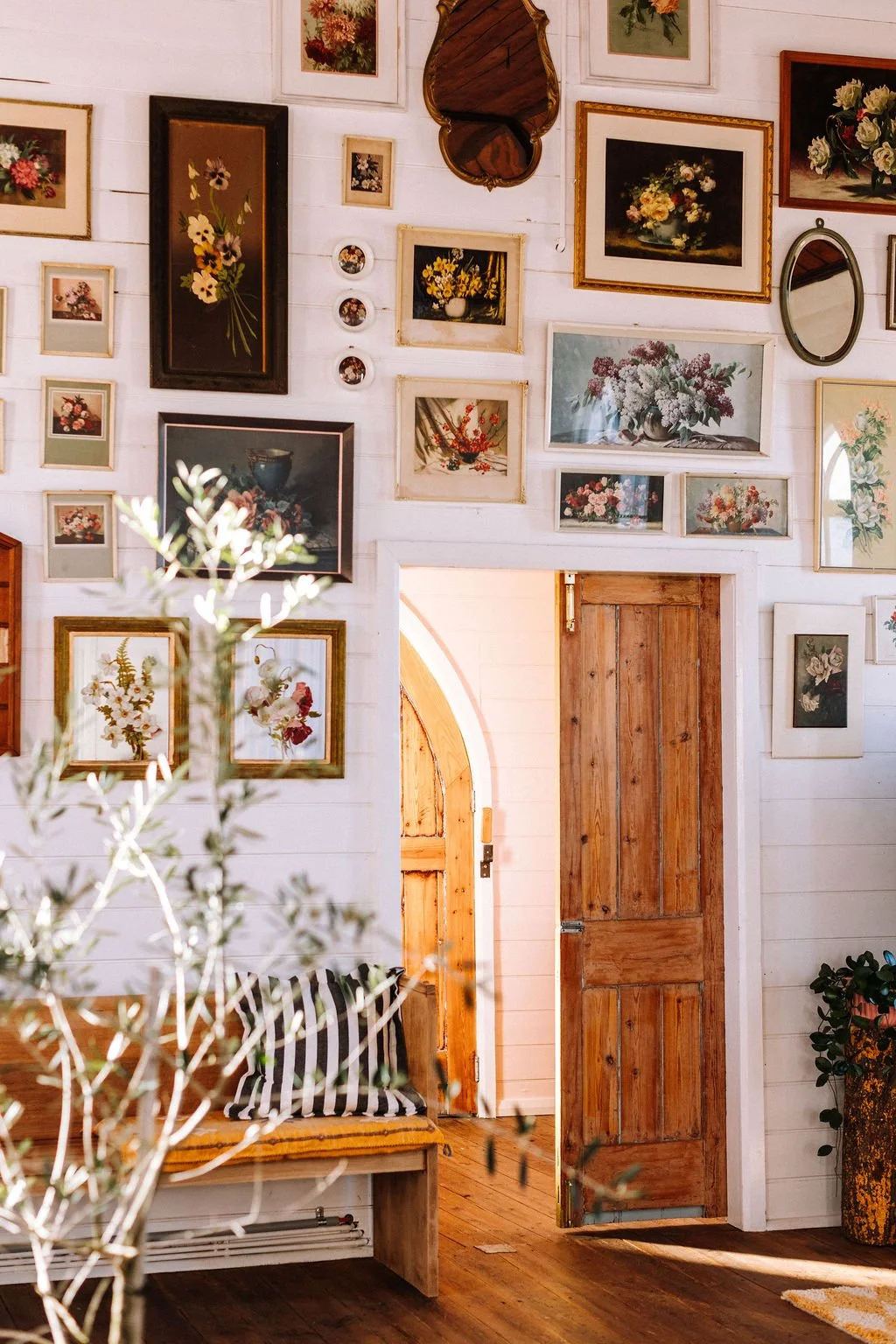 Interior of a room with white wooden walls decorated with numerous framed floral art prints and photographs. A wooden door is open, revealing a glimpse of another room. There is a bench with striped pillow, a plant in a vase, and a small mirror on th