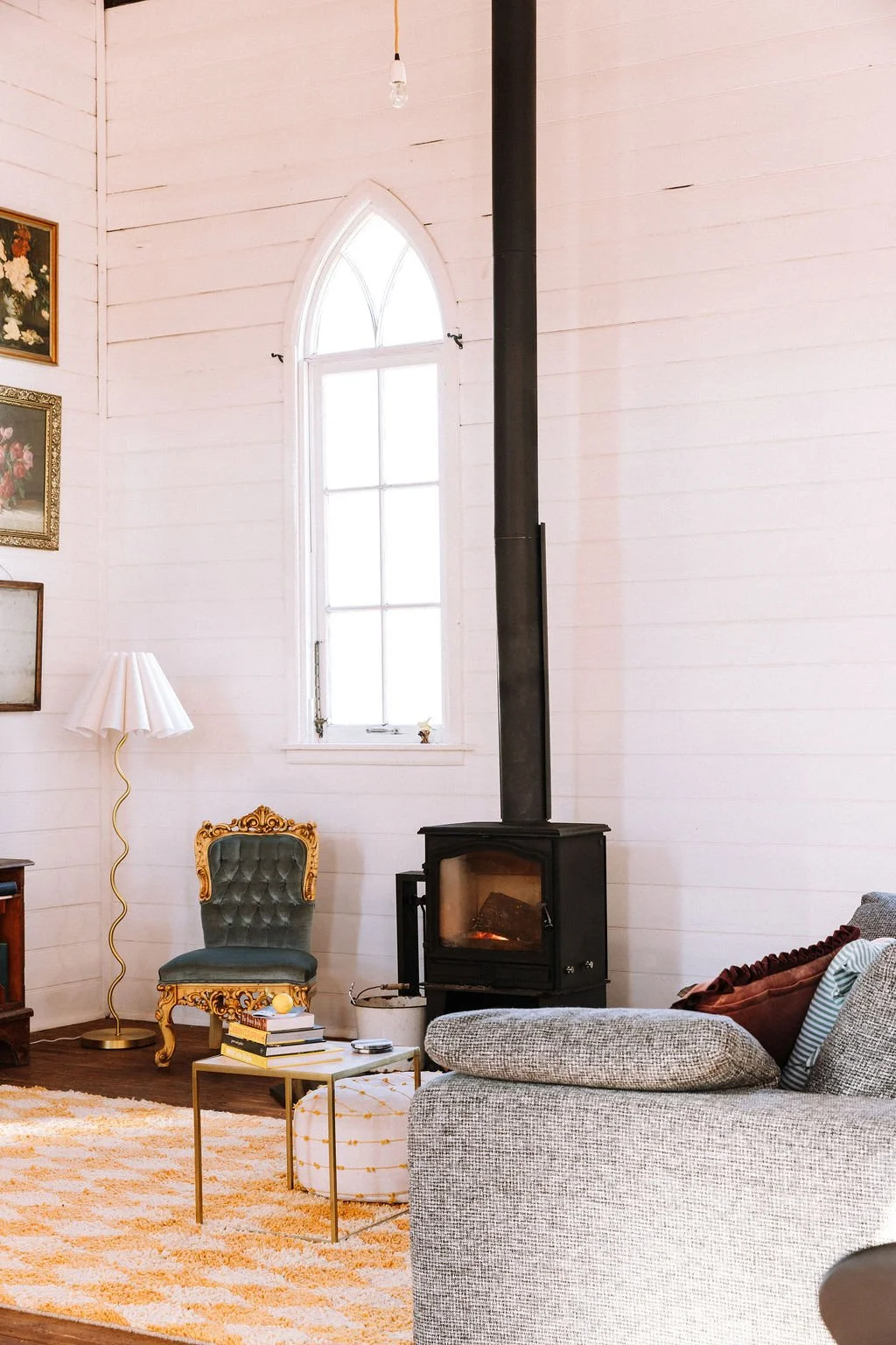 Cozy living room with white wooden walls, a black wood stove with a pipe, a large arched window, a vintage green and gold armchair, a white lamp with a wavy gold base, a small white table with books and a round ottoman, and framed floral artwork on t
