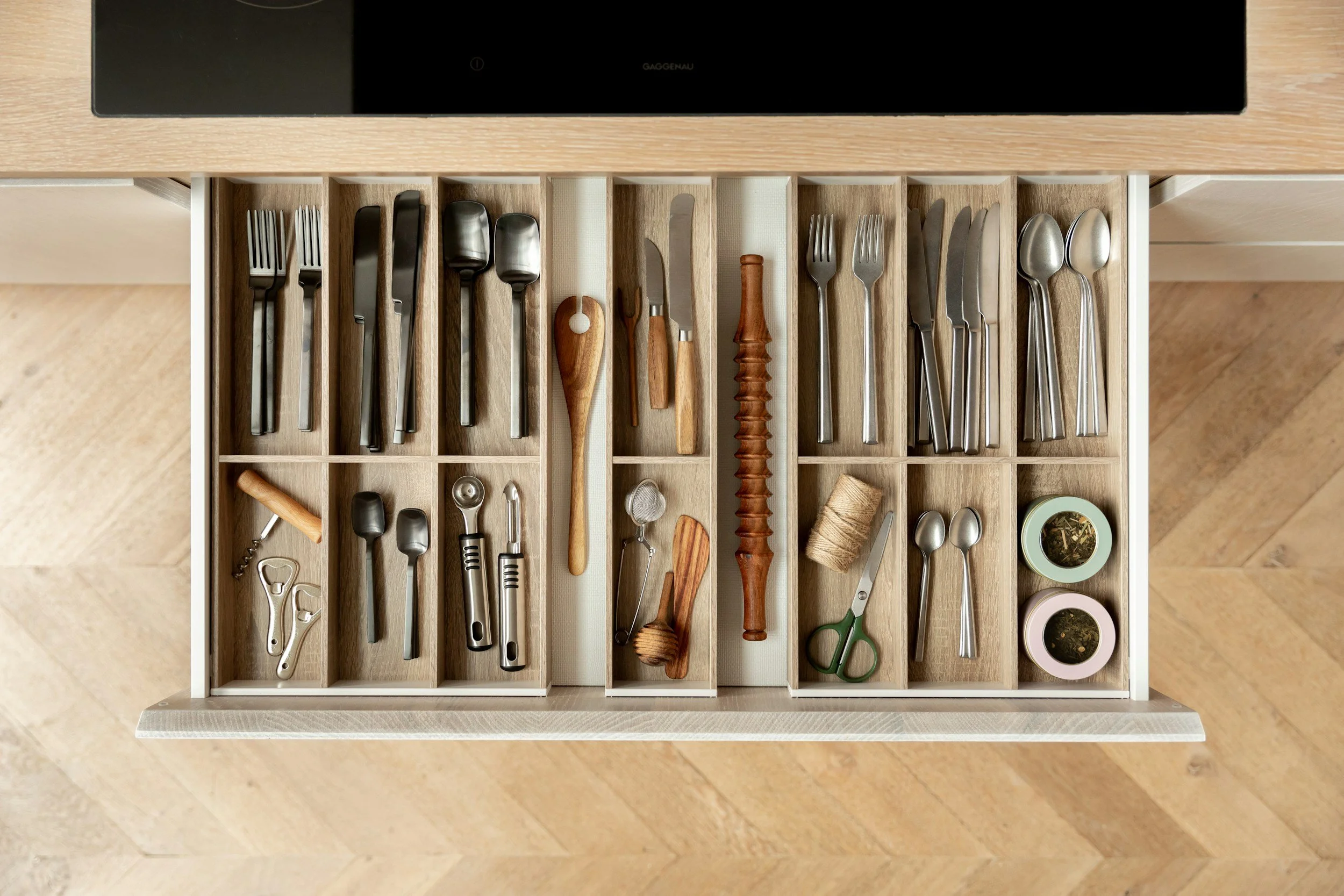 Drawer containing various kitchen utensils such as forks, knives, spoons, a corkscrew, scissors, and small containers for herbs.