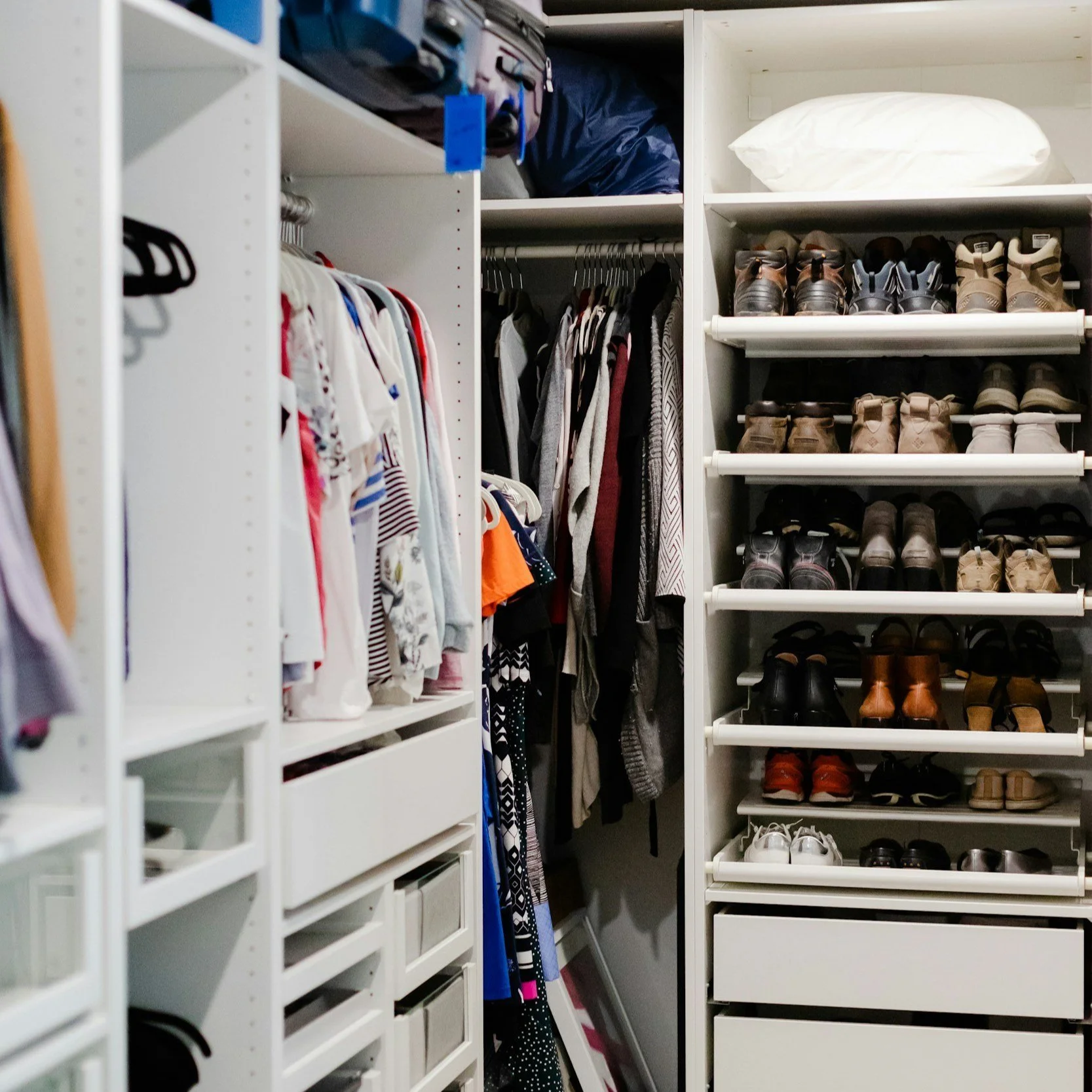 Organized walk-in closet with clothing on hangers, shoes on white shelves, and storage drawers.
