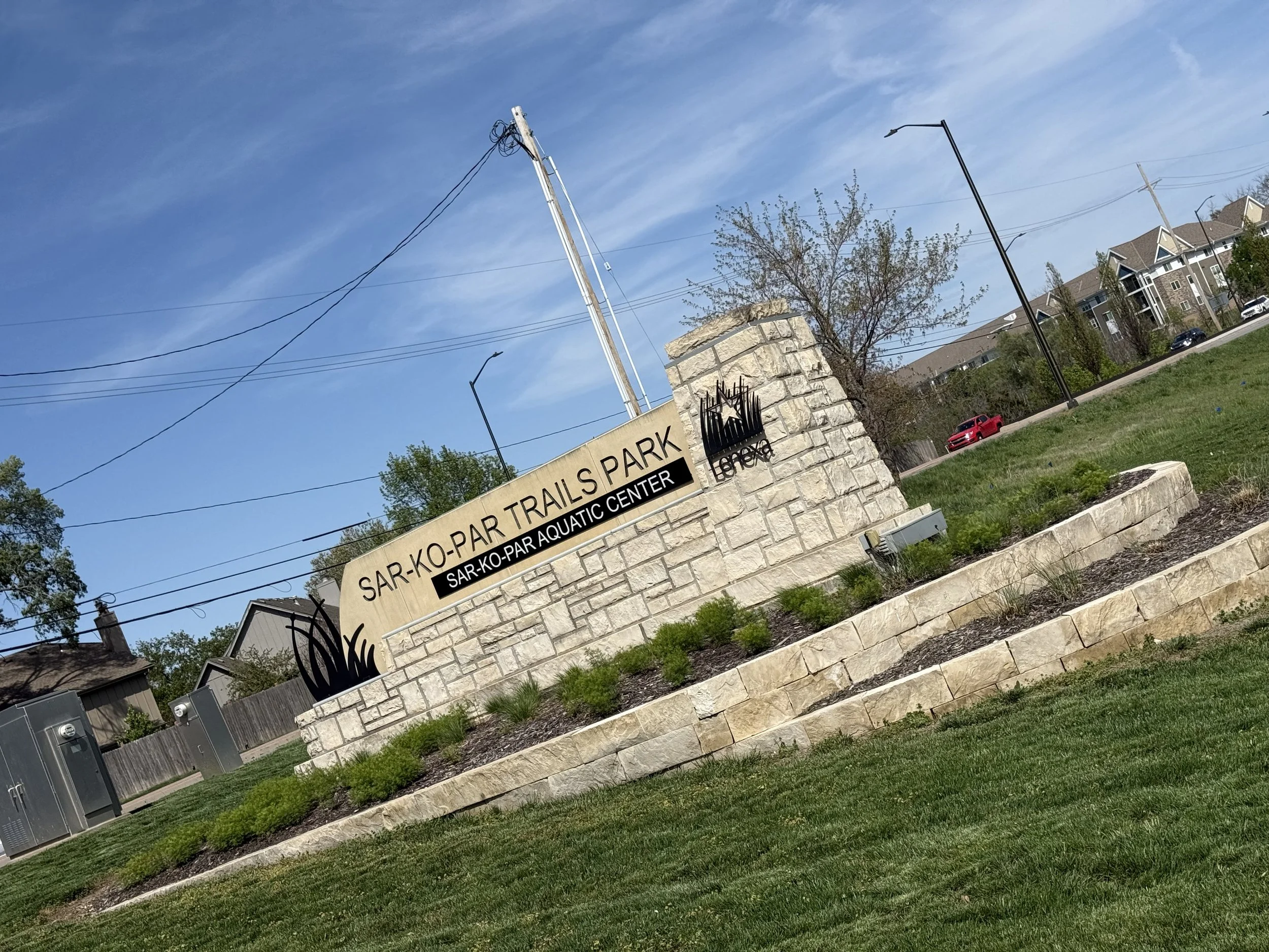 Sign for Sar-Ko-Par Trails Park and Sar-Ko-Par Aquatic Center, with landscaped area in front, trees, power lines, and buildings in the background under a blue sky.