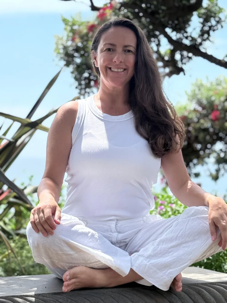 Daiana, with long dark hair, sitting cross-legged outdoors on a log and smiling, with trees and flowering plants behind her