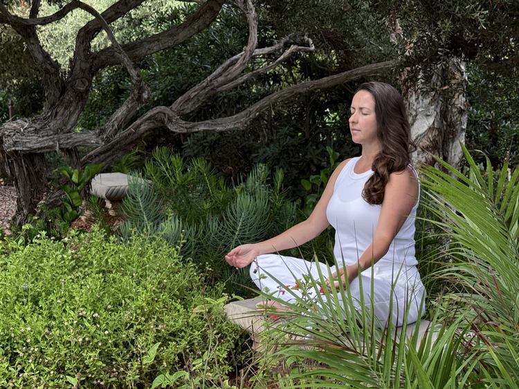 Daiana meditating outdoors in a lush garden, sitting cross-legged with eyes closed and hands resting on her knees