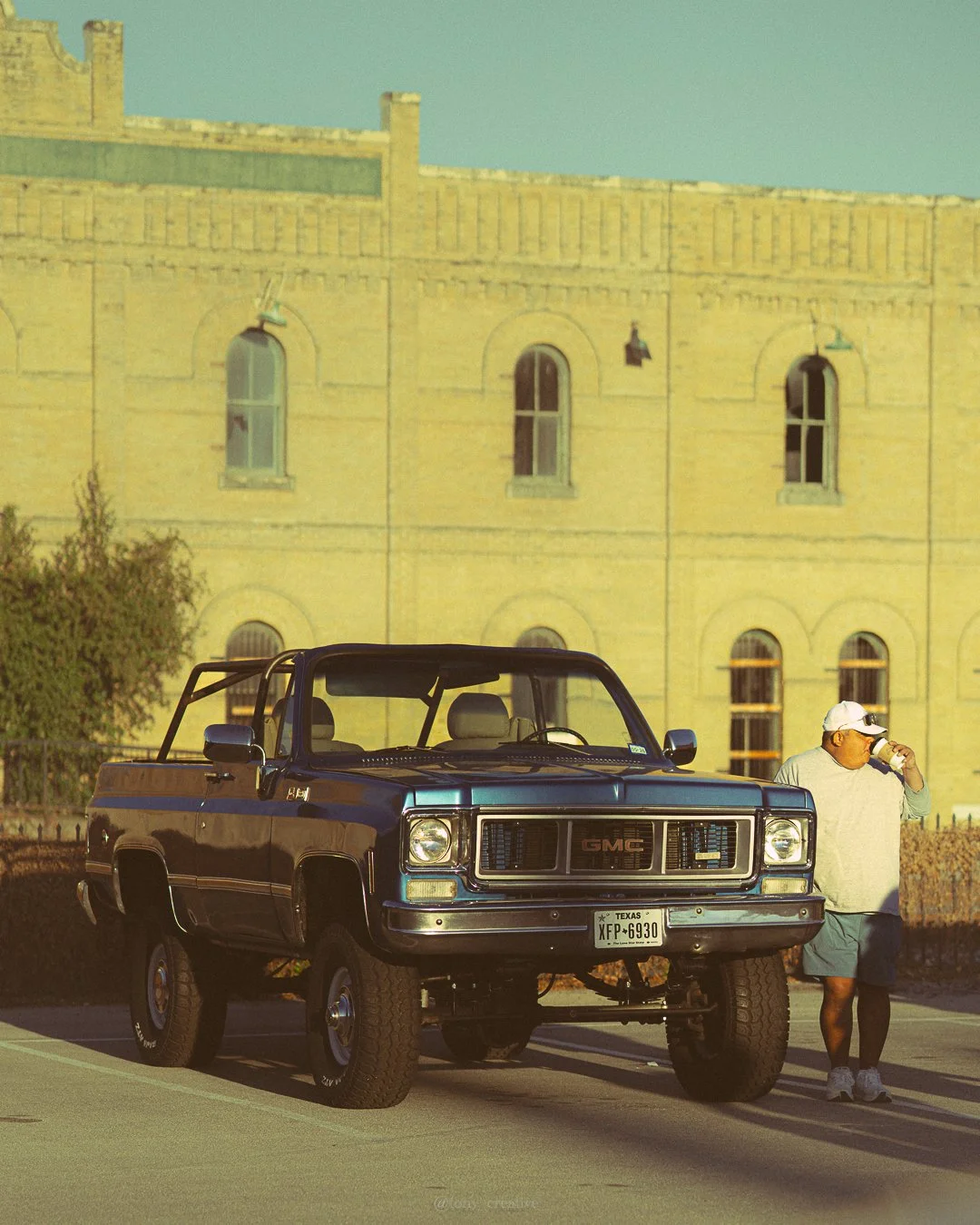 A black GMC pickup truck parked on the street, with a man standing nearby drinking from a cup, in front of a yellow brick building with arched windows, under a clear sky.