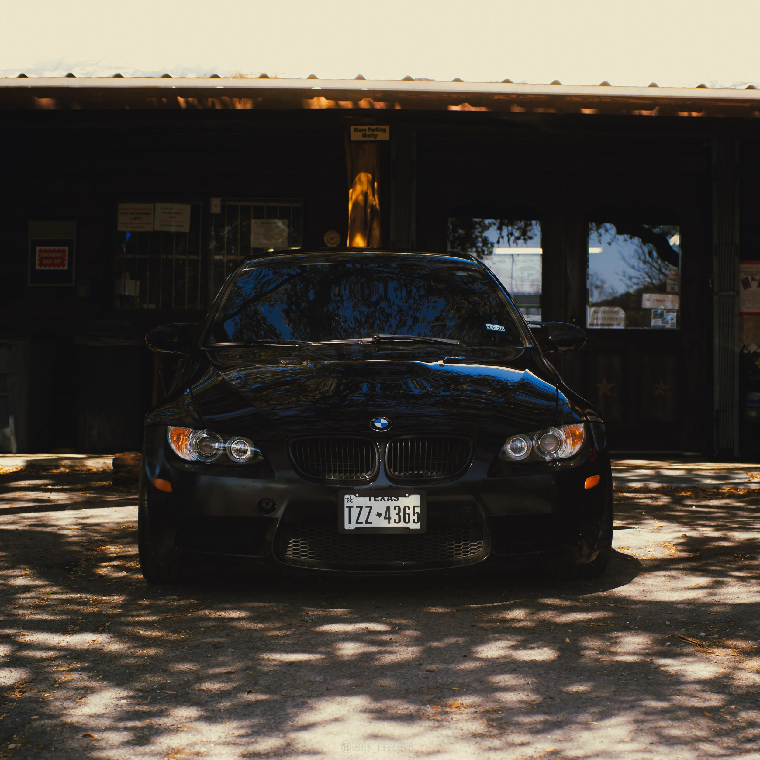 A black BMW car parked in front of a dark building with windows and a sign that reads 'Store Parking Only'. Shadows and trees are reflected on the car's surface.