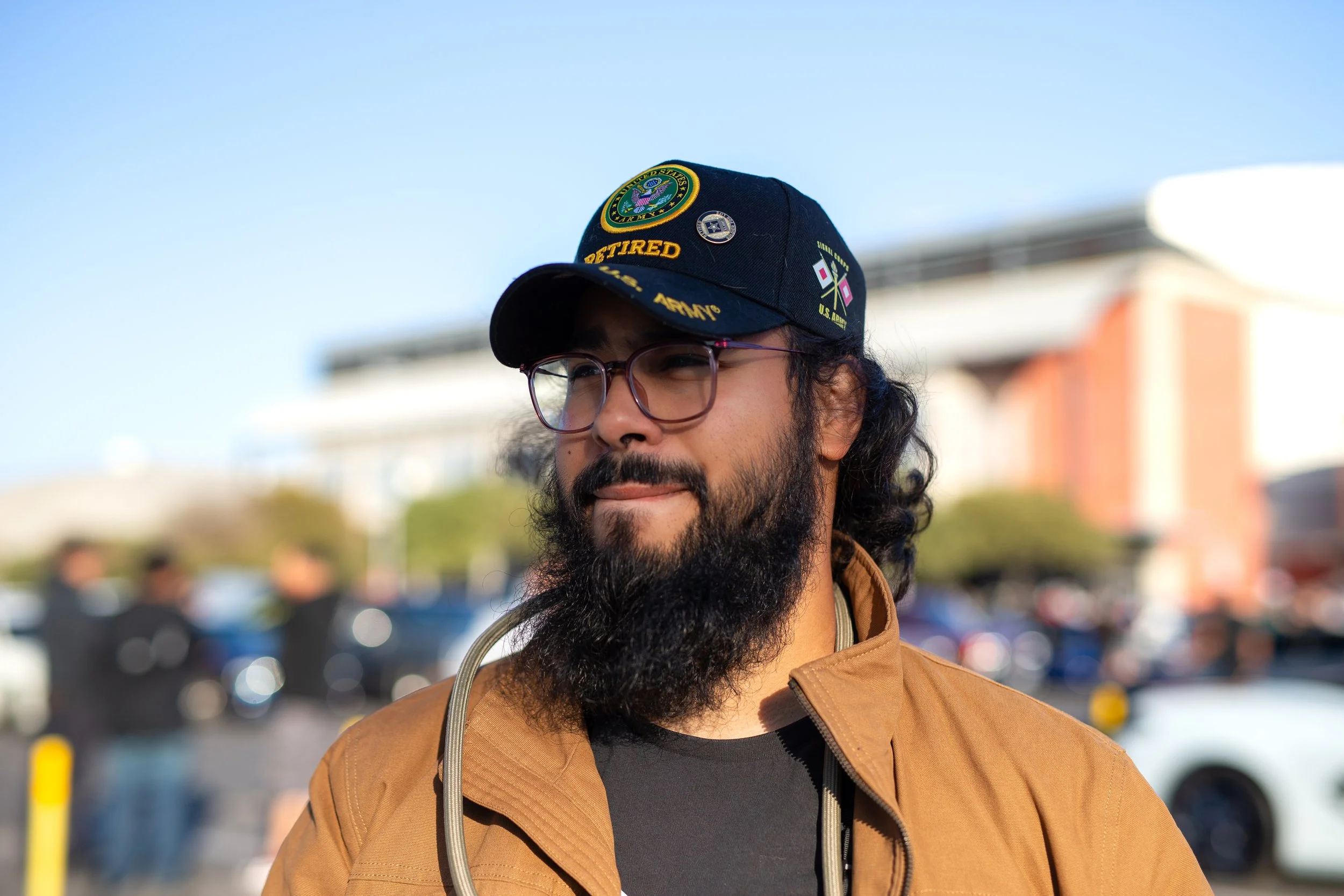 A man with glasses and a beard wearing a navy military cap with patches, including one that says "Retired" and "U.S. Army," standing outdoors with a background of a parking lot and city buildings.