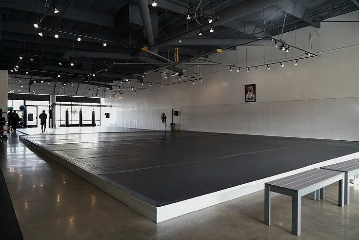 Empty martial arts training room with black mats, white walls, benches, and large windows at the front.