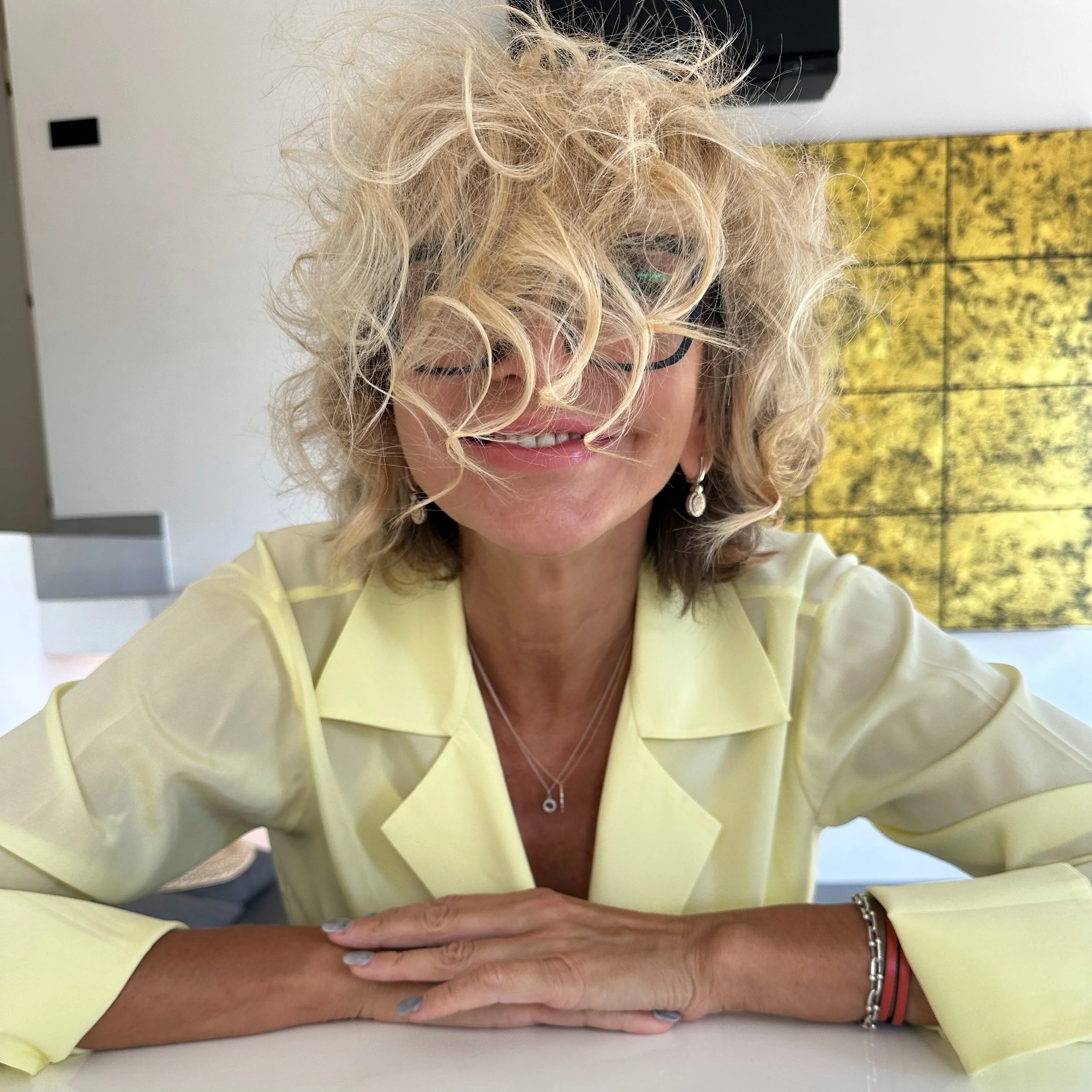 Artist Milla Petrova, with curly blonde hair, glasses, and jewelry, smiles while sitting at a table in a bright room with a yellow, textured piece of art on the wall behind her.