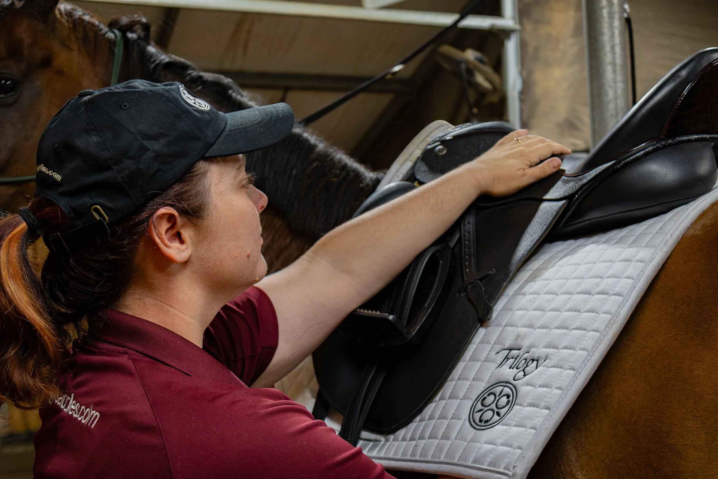 A woman in a maroon shirt and black cap placing her hand on a horse's saddle to check the saddle's fit on the horses back.