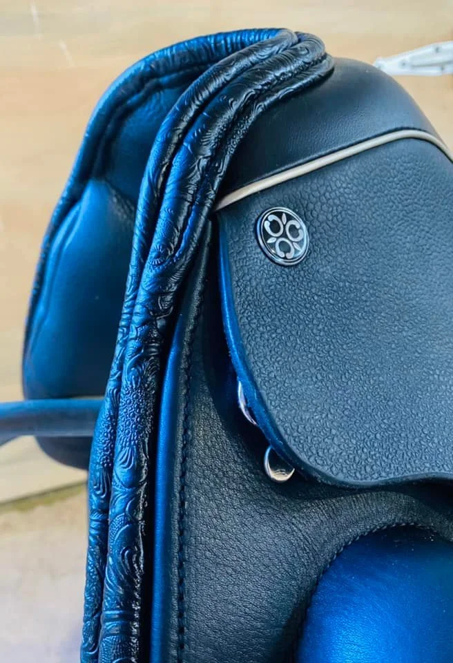 Close-up of a black leather saddle with intricate embossed details and a decorative metal emblem.