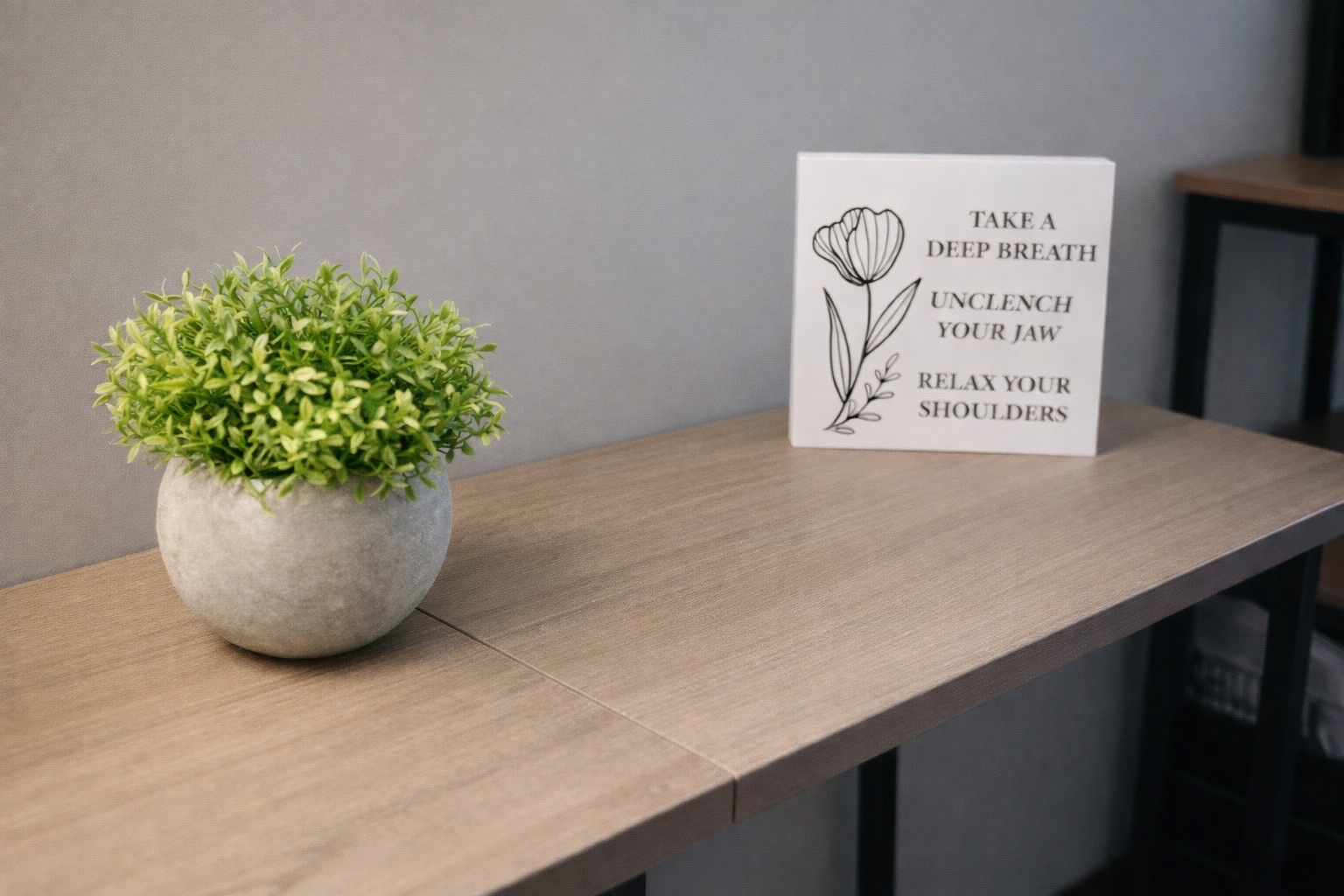A potted green plant and a white sign with a black outline flower drawing and the text 'Take a deep breath, unclench your jaw, relax your shoulders' on a wooden table against a gray wall.