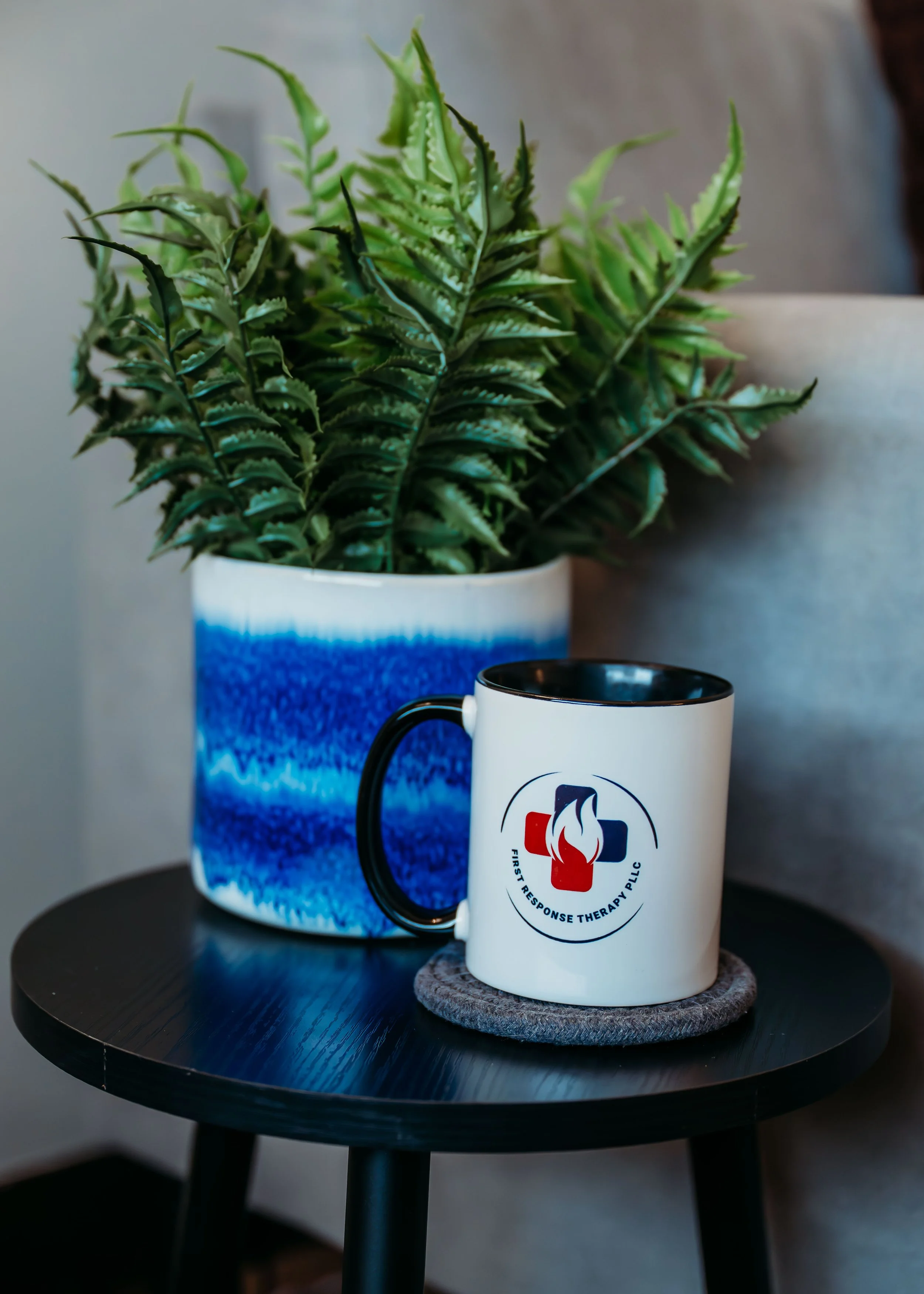 A white mug with a First Response Therapy logo sitting on a gray felt coaster on a small black table, with a large green fern in a blue-and-white ceramic pot nearby.