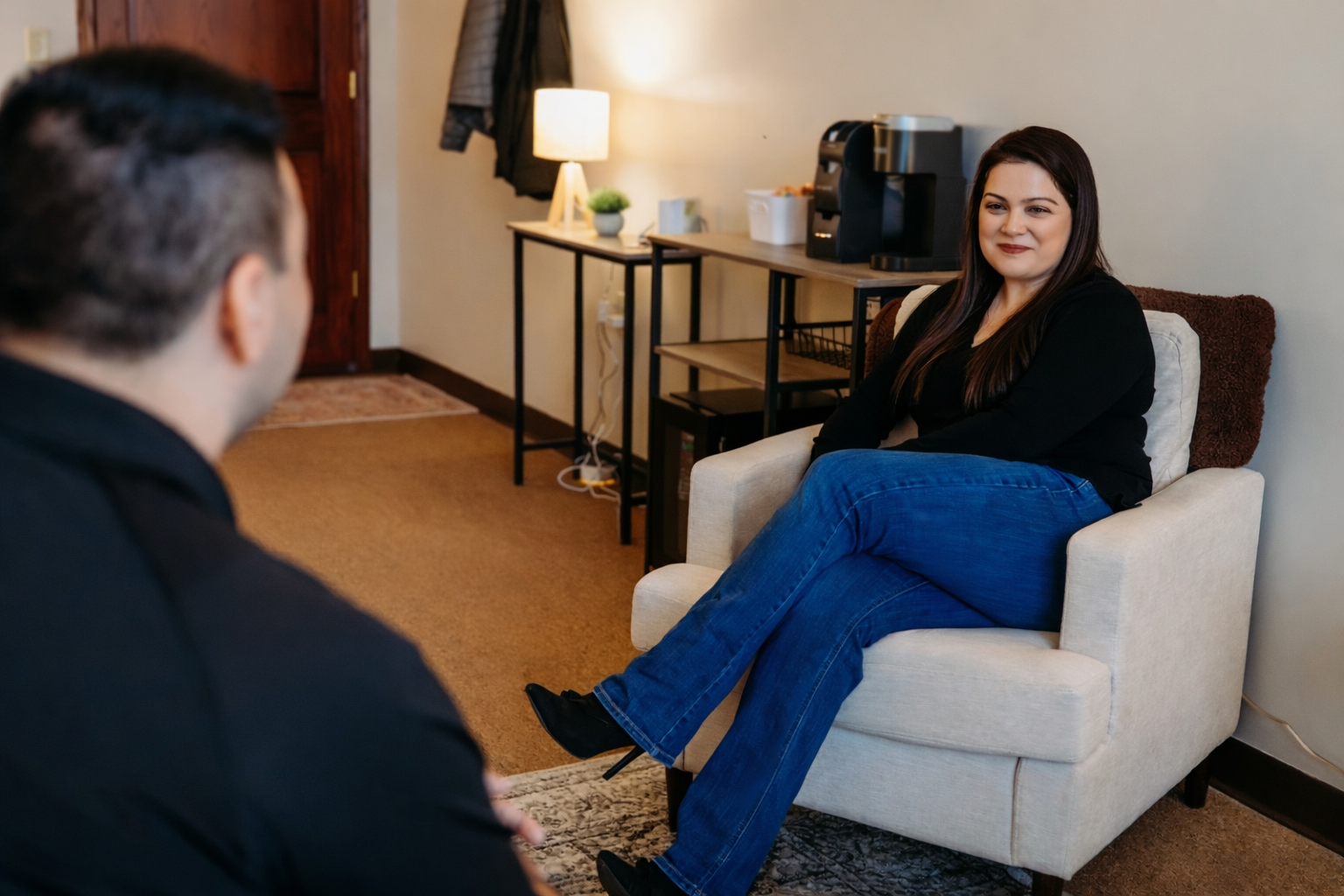 A woman sitting on a white armchair, smiling, with her legs crossed, engaging in conversation with a man whose back is to the camera. The setting appears to be a cozy living room or office area with a side table, a table lamp, and a coffee maker in the background.