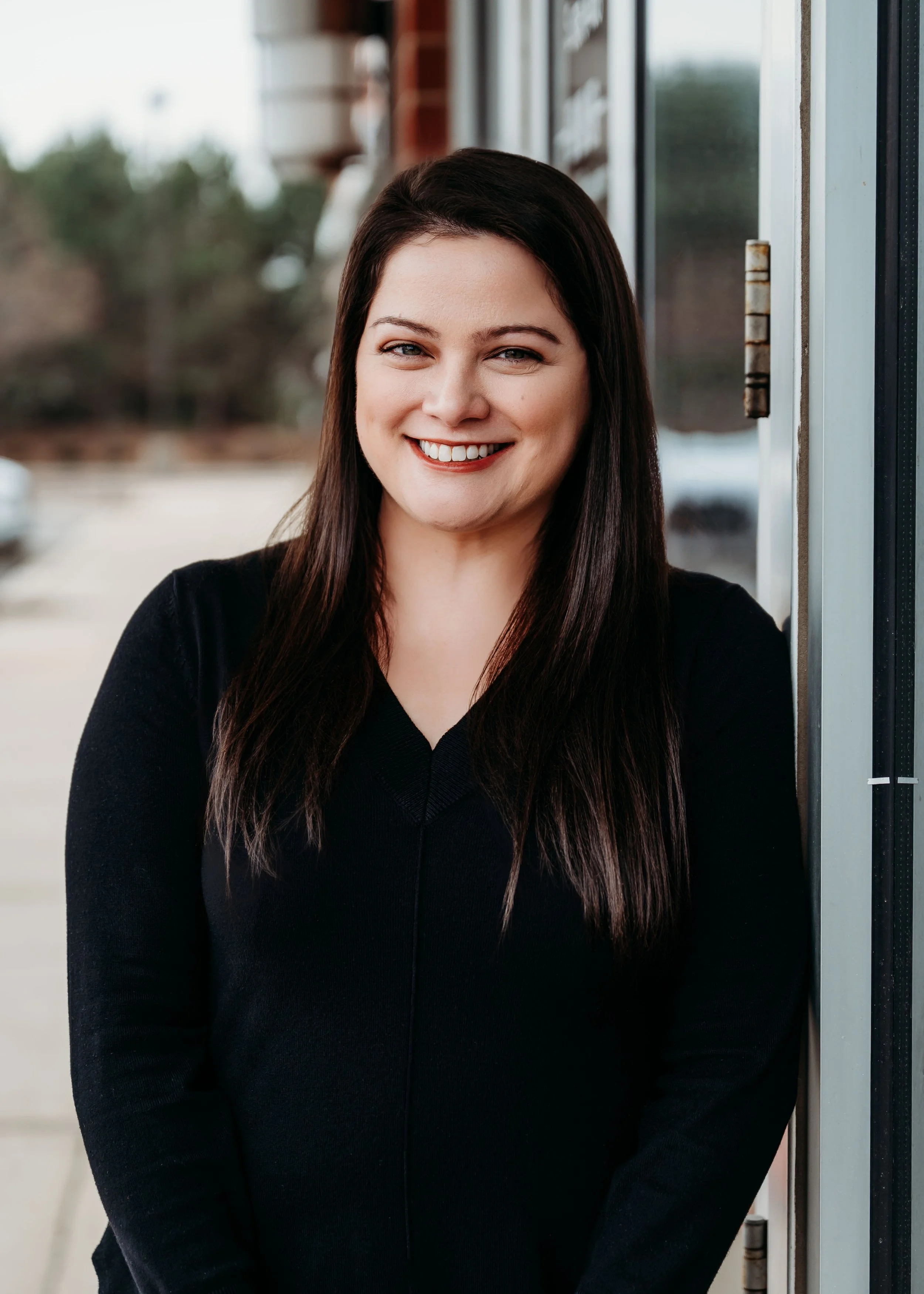 A woman therapist with long dark hair and a black top smiling outdoors in front of a storefront window.