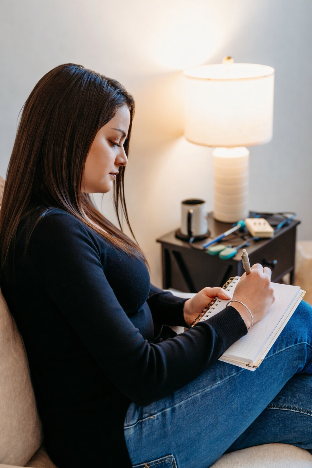 A woman with long brown hair sitting on a beige couch, writing in a notebook with a pen, beside a side table with a lamp, mug, and assorted tools.