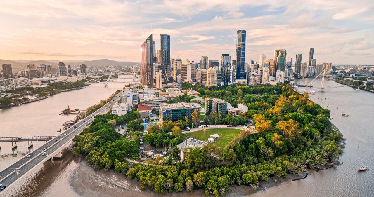 Aerial view of Brisbane skyline with tall modern buildings, river, and green parklands, during sunset.