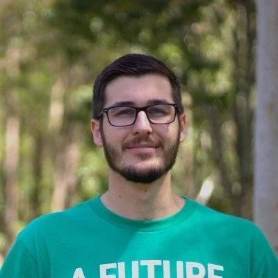 A young man with glasses and a beard, wearing a teal shirt, standing outdoors in a park.