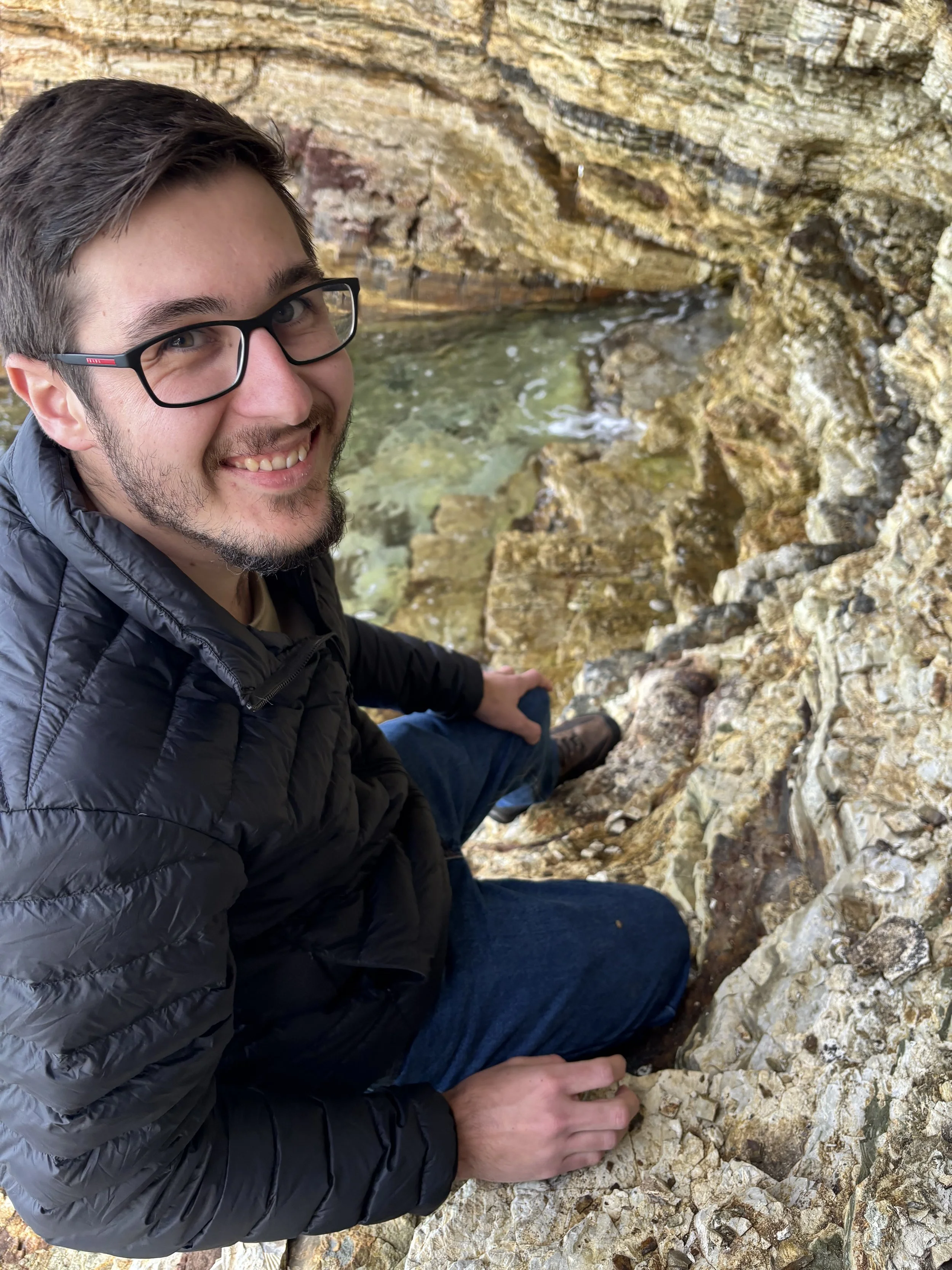 A man with glasses and a beard, smiling, is sitting on rocks near a small body of water inside a rocky cave or crevice.