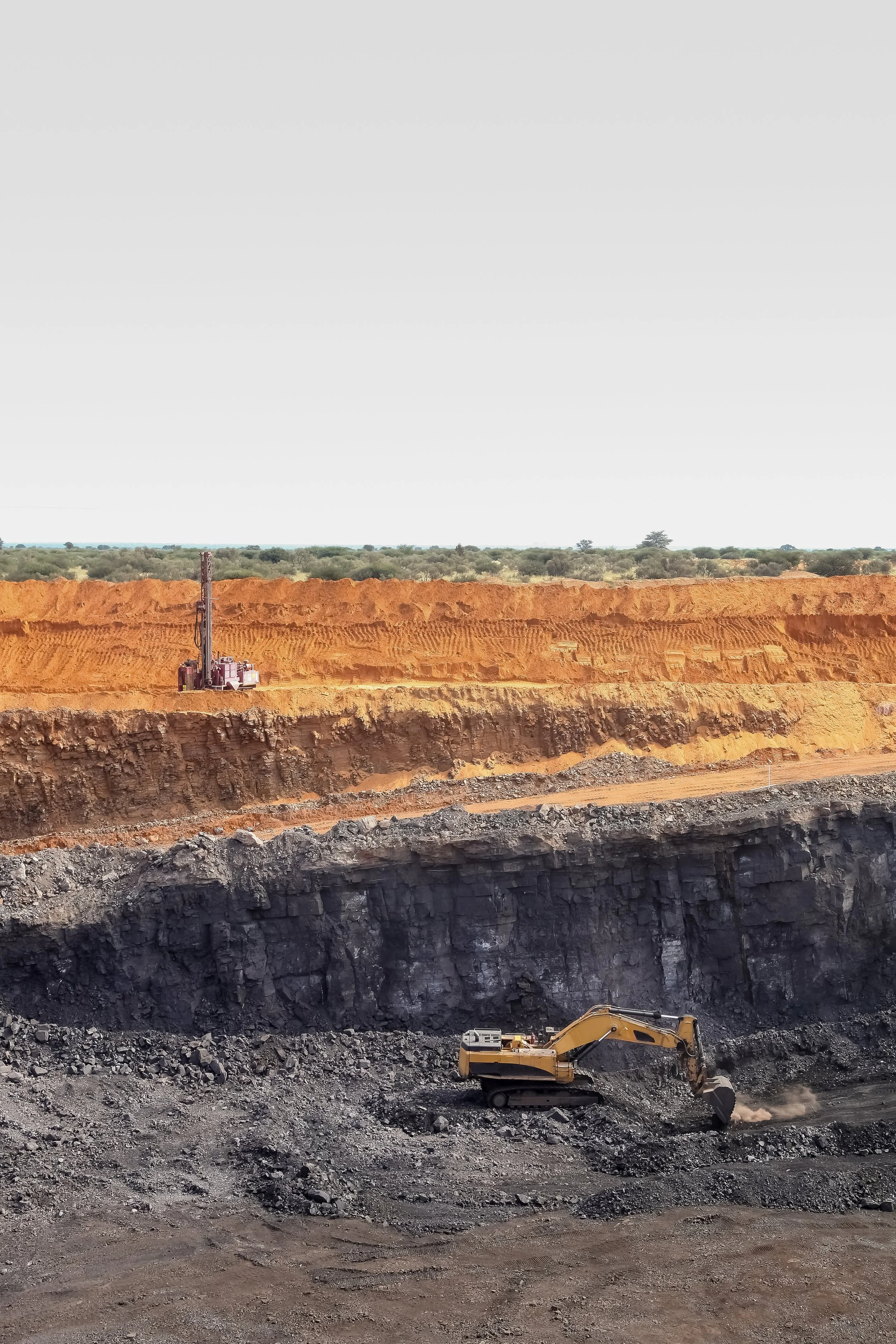 Image of an open-pit mining operation with excavators working in a large dark coal or mineral extraction site, with layers of earth and rock visible, and a flat, distant landscape in the background.