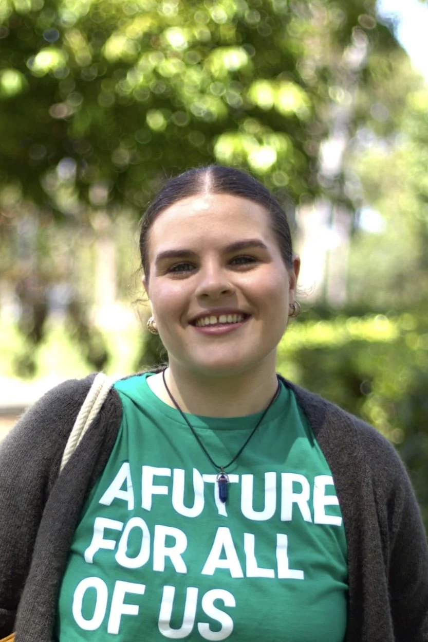 A young woman outdoors smiling, wearing a green T-shirt with the words 'FUTURE FOR ALL OF US', a grey cardigan, and a necklace with a dark-colored pendant.