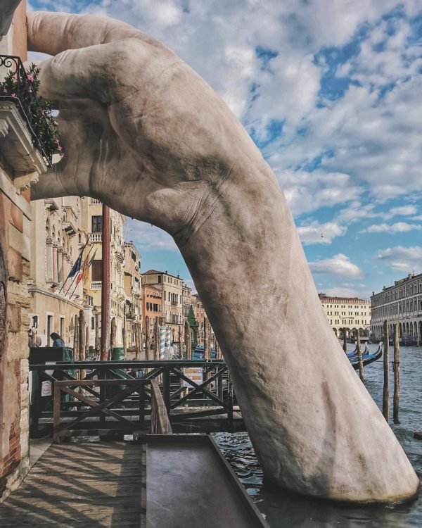 Large hand sculpture partially submerged in water along a canal, with historic European-style buildings and a cloudy sky in the background.