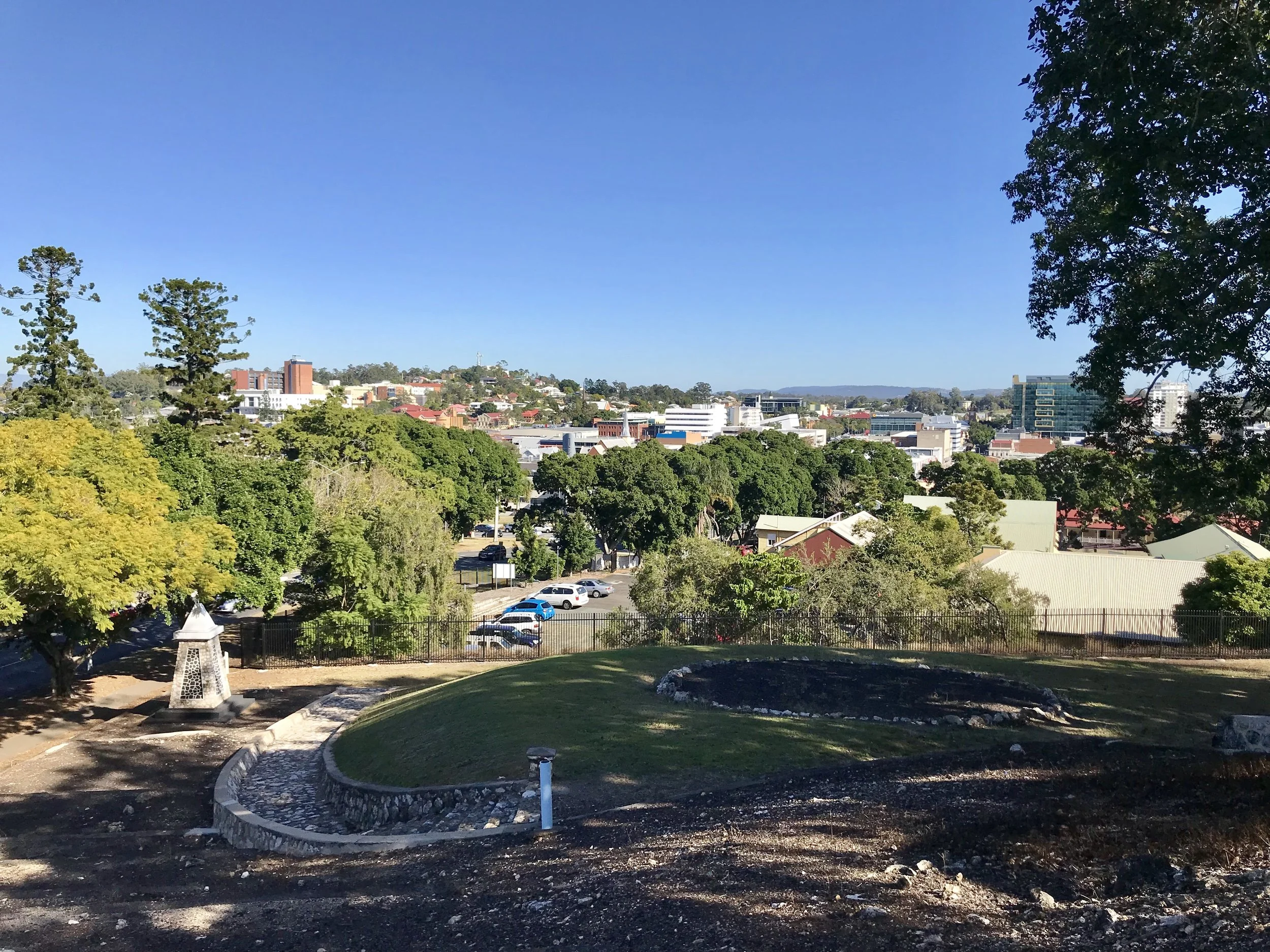 A cityscape view with trees, buildings, and a parking lot under a clear blue sky, taken from an elevated location.