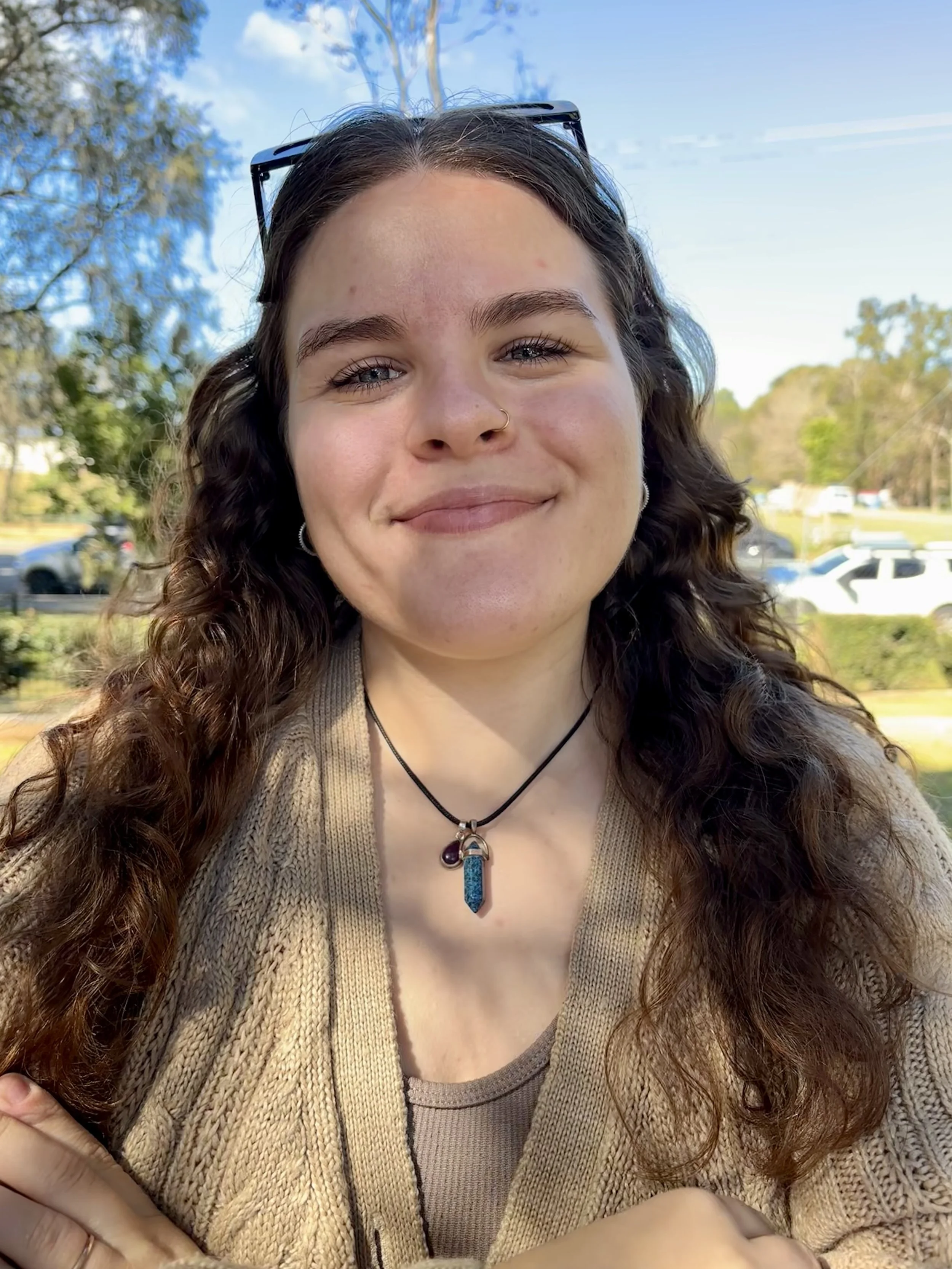 Close-up of a young woman with long curly hair, sunglasses on her head, wearing a tan cardigan, a necklace with a blue pendant, smiling outdoors on a sunny day with trees and parked cars in the background.