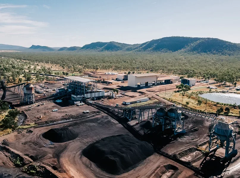 An aerial view of an industrial mining site with conveyor belts, large piles of extracted material, industrial buildings, parked vehicles, a pond, and mountains in the background.
