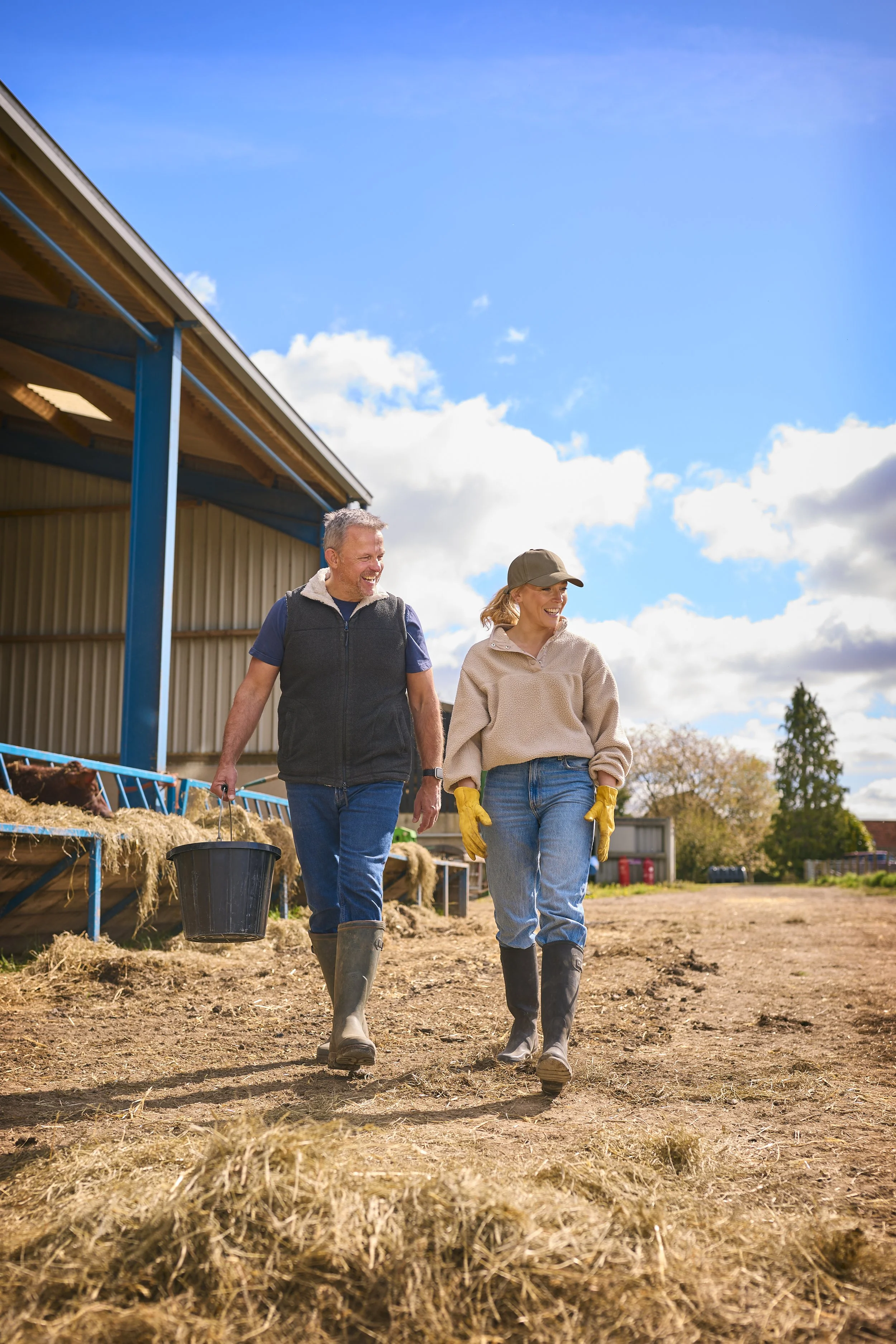 A man and woman walking on a farm, smiling and enjoying their work. The woman is wearing yellow gloves and a cap, while the man is holding a black bucket. They are outdoors near a barn with straw and farm animals visible. The sky is partly cloudy and bright.