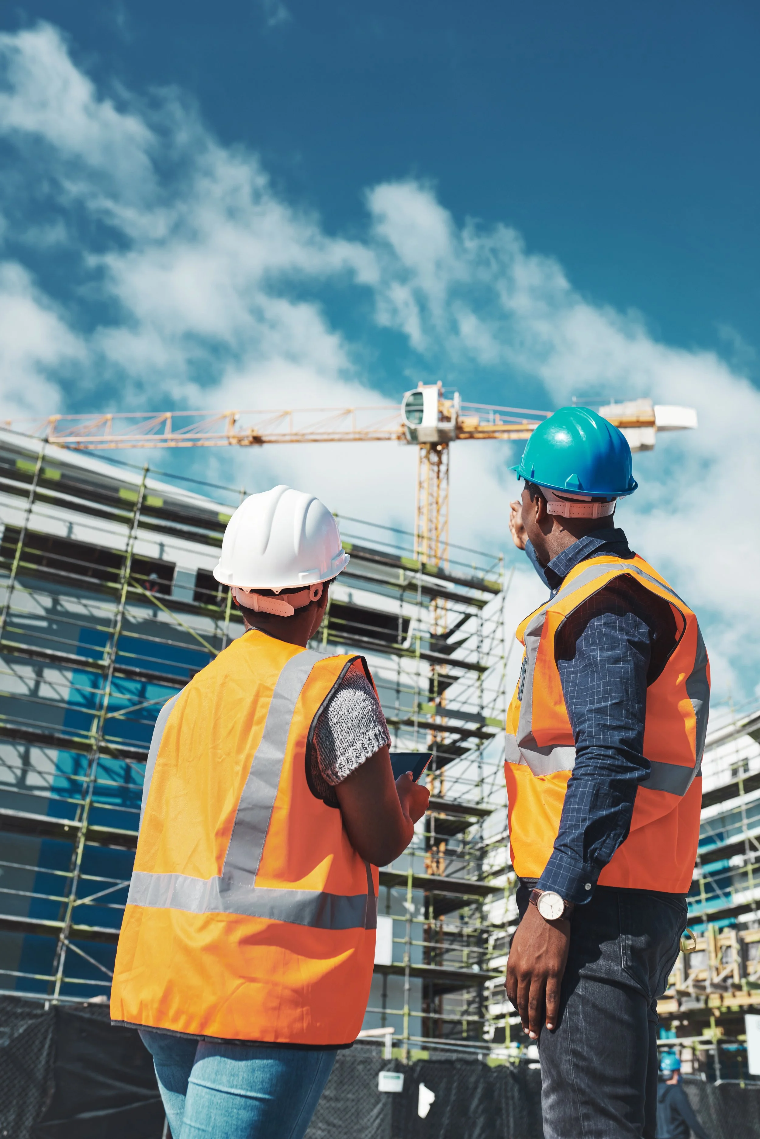 Two construction workers wearing safety helmets and vests looking at a construction site with scaffolding and a crane under a partly cloudy sky.