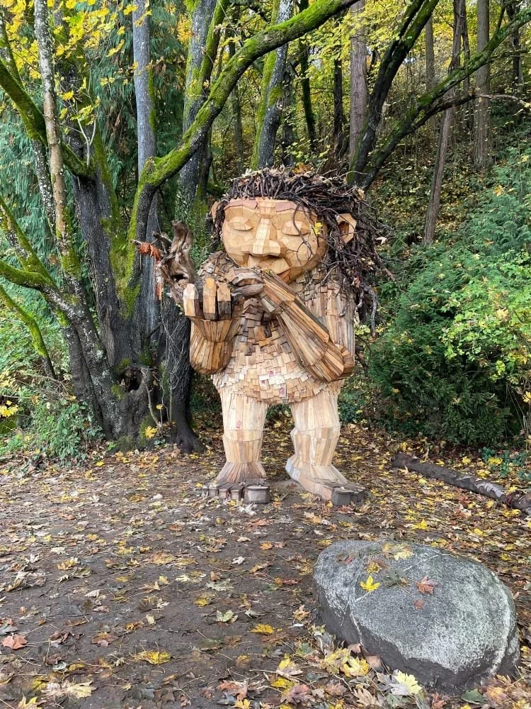 Large wooden sculpture of a person with long, curly hair, holding a flute, standing in a wooded area surrounded by autumn leaves and trees.