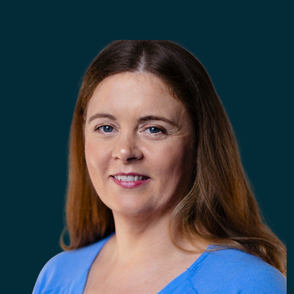 A woman with long brown hair wearing a blue top, smiling at the camera against a plain background.