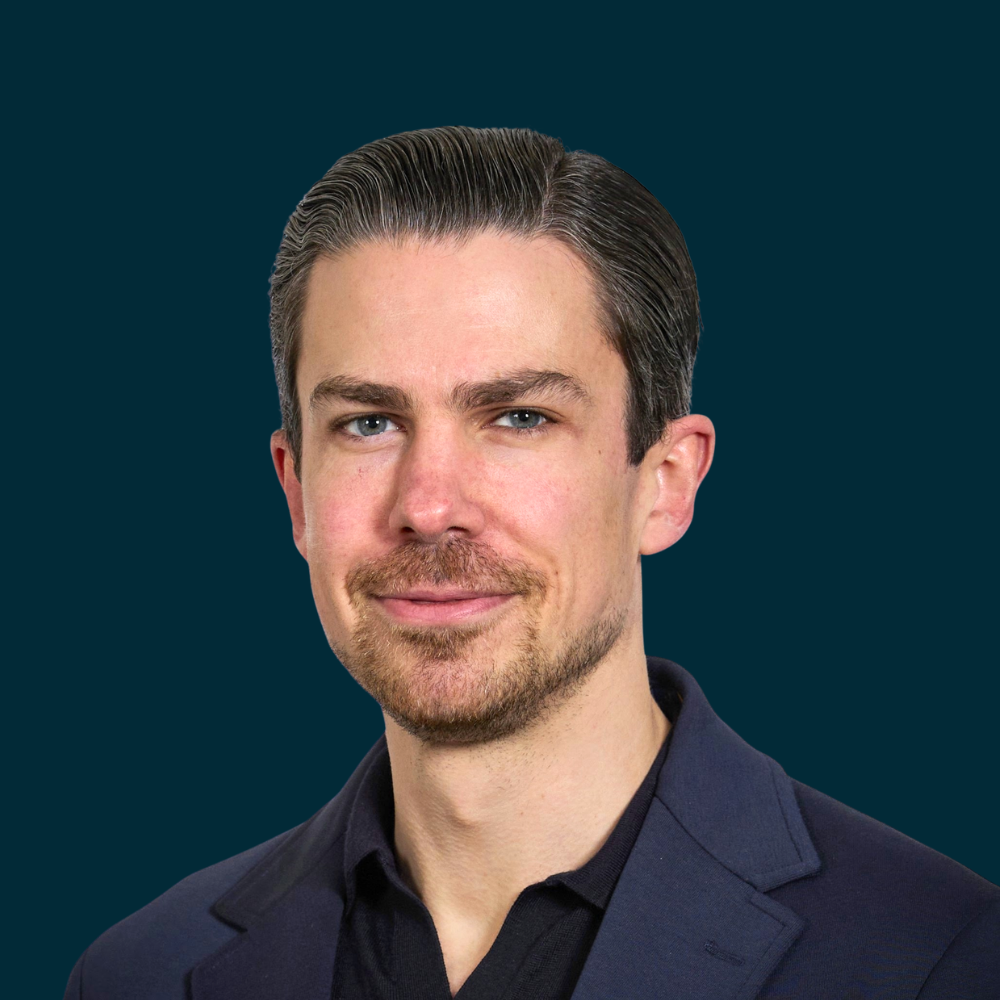A portrait of a man with dark hair and stubble, wearing a dark suit jacket and a dark shirt, looking at the camera.