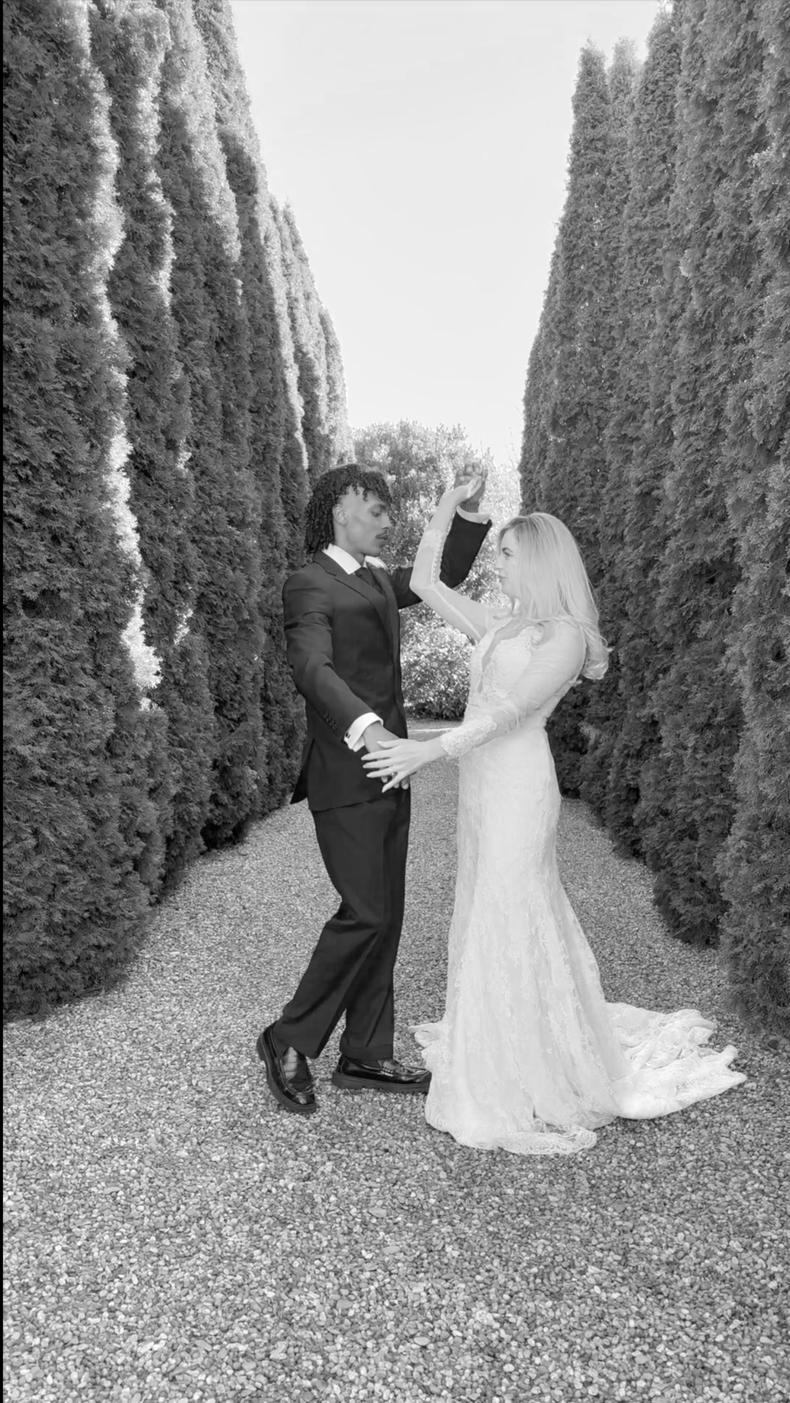 A married couple, a man in a suit and a woman in a wedding dress, dancing together in an outdoor garden with tall, closely lined trees.
