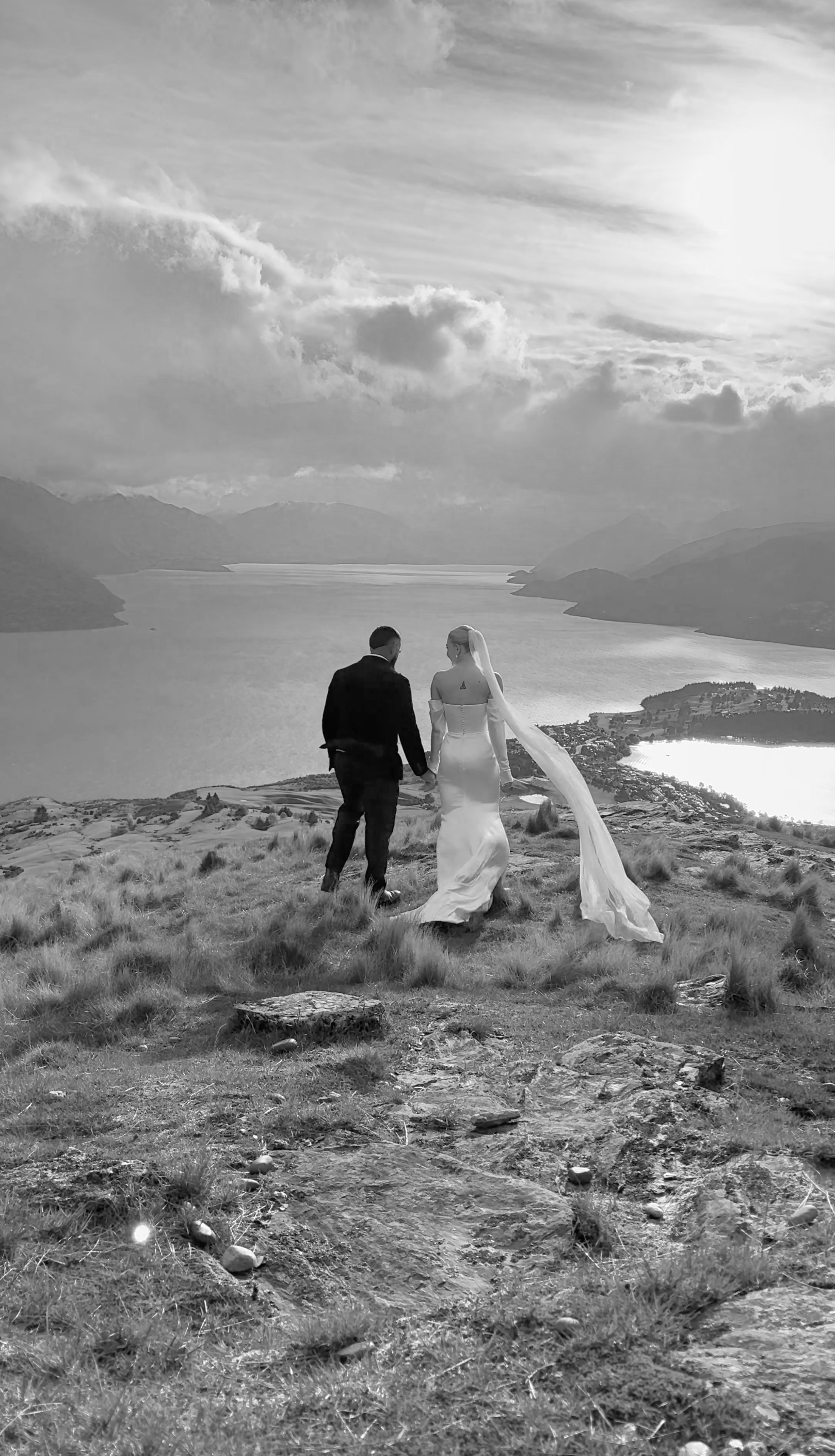 A black and white photo of a couple in wedding attire holding hands and walking on a grassy hill with a lake and mountains in the background.