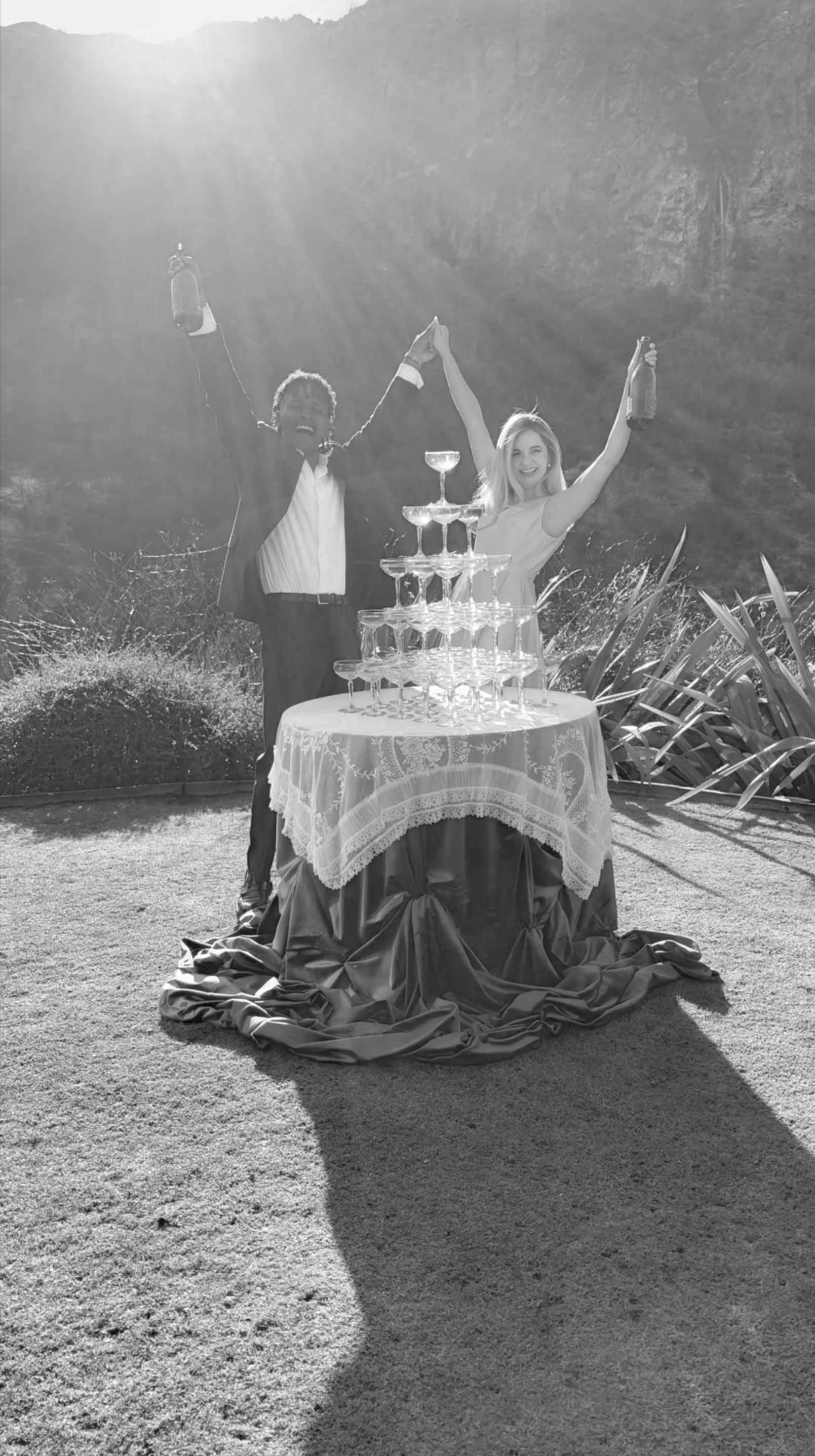 A man and woman celebrating outdoors with a champagne tower, holding bottles, smiling, with sunlight in the background.