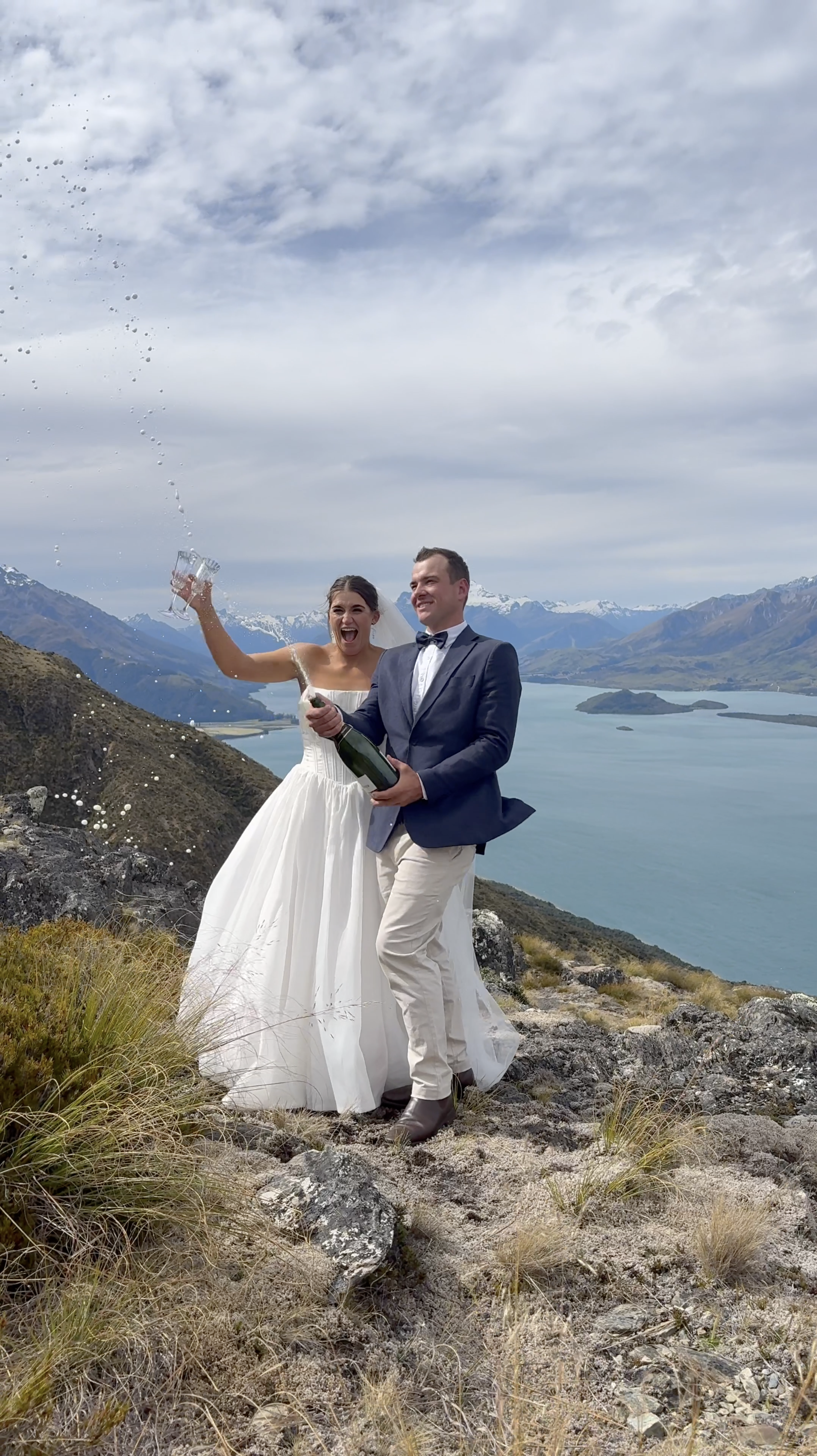 Bride and groom celebrating on a mountain with a lake and snow-capped mountains in the background, snow is being poured into champagne glasses.