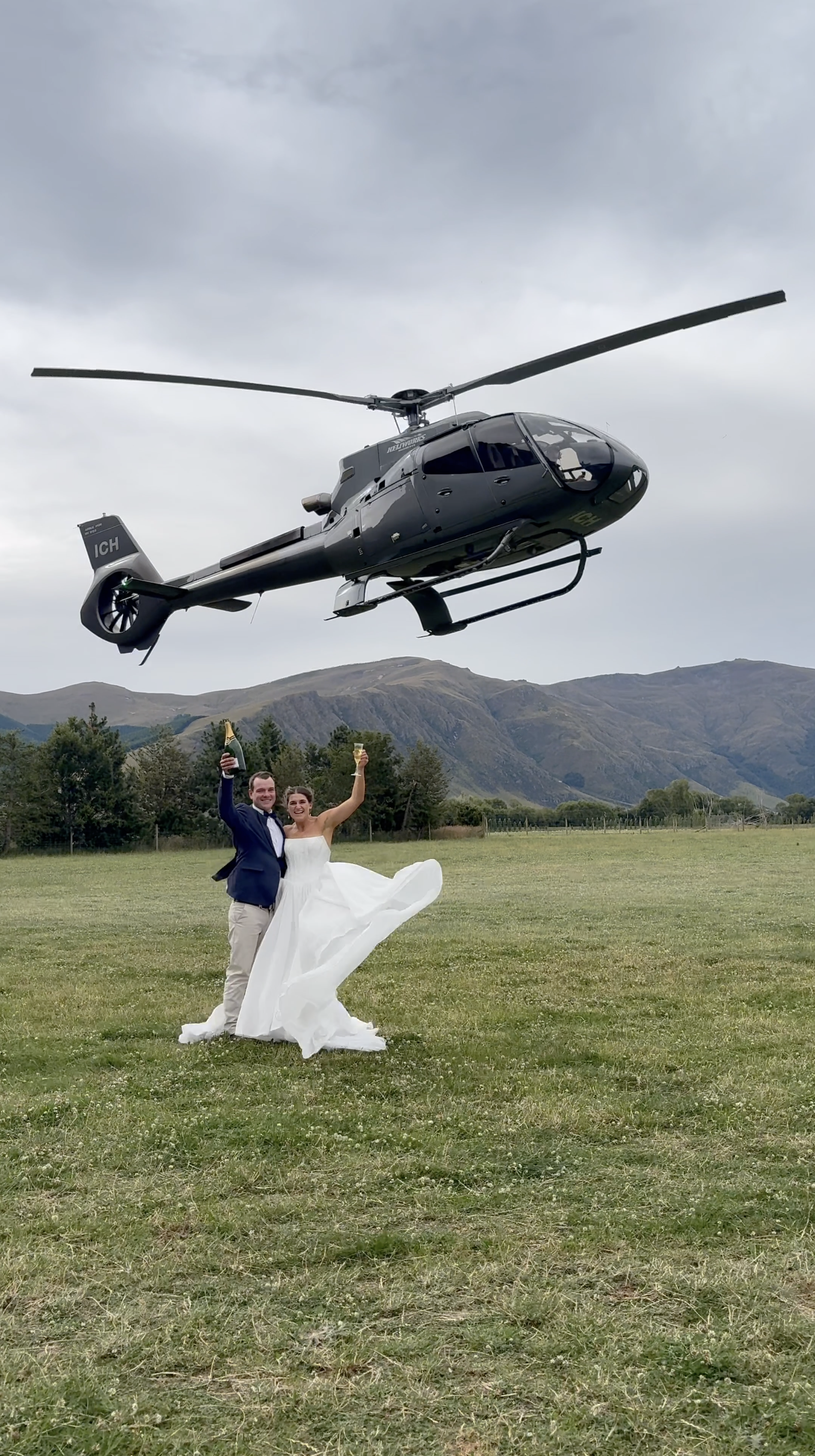 A couple in wedding attire celebrating with drinks in hand while standing on grass during the daytime, with a helicopter flying overhead and mountains in the background.
