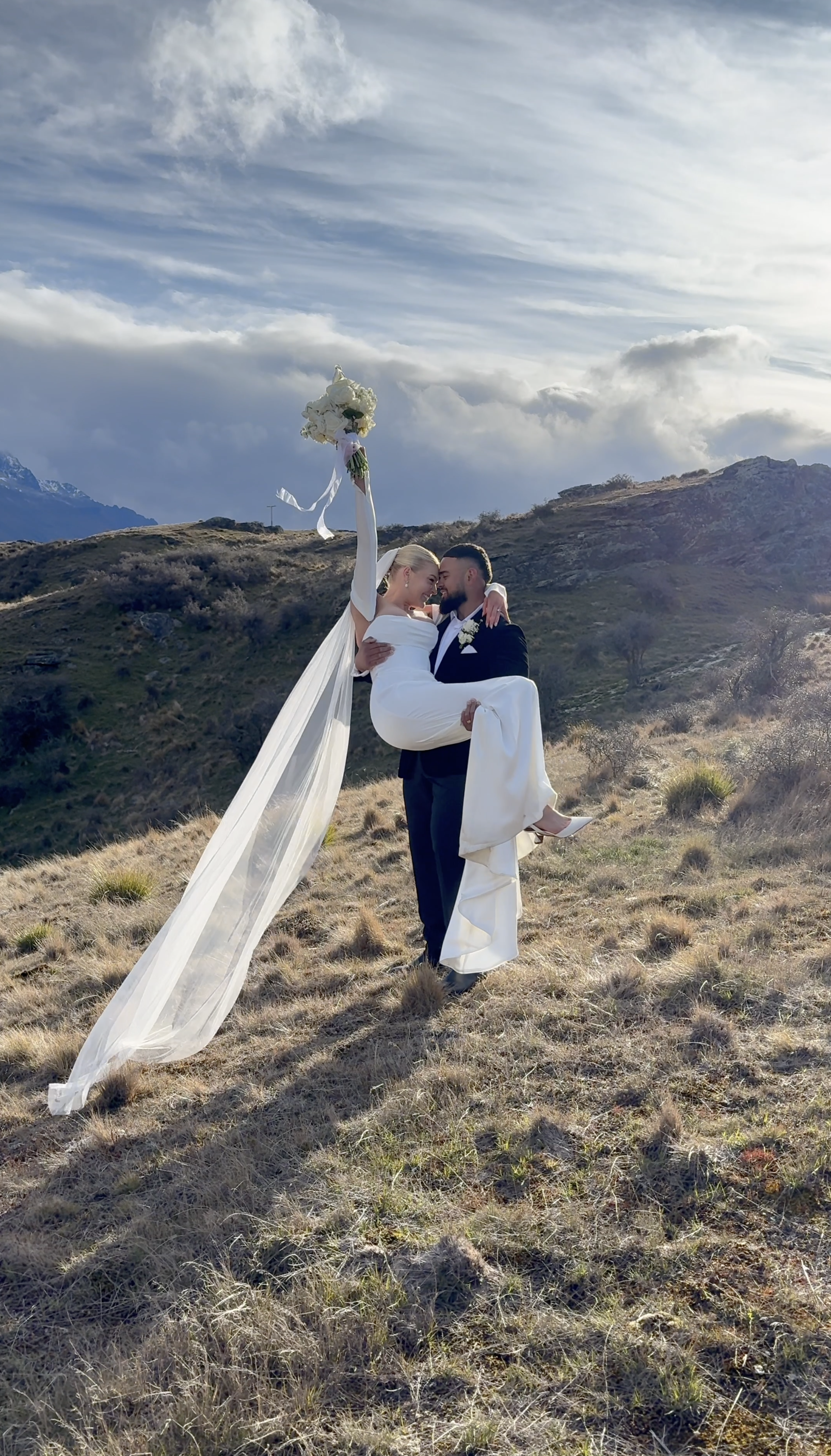 A newlywed couple in wedding attire, with the groom carrying the bride, stands on a grassy hillside with mountains and a cloudy sky in the background. The bride holds a bouquet of white flowers raised in the air.