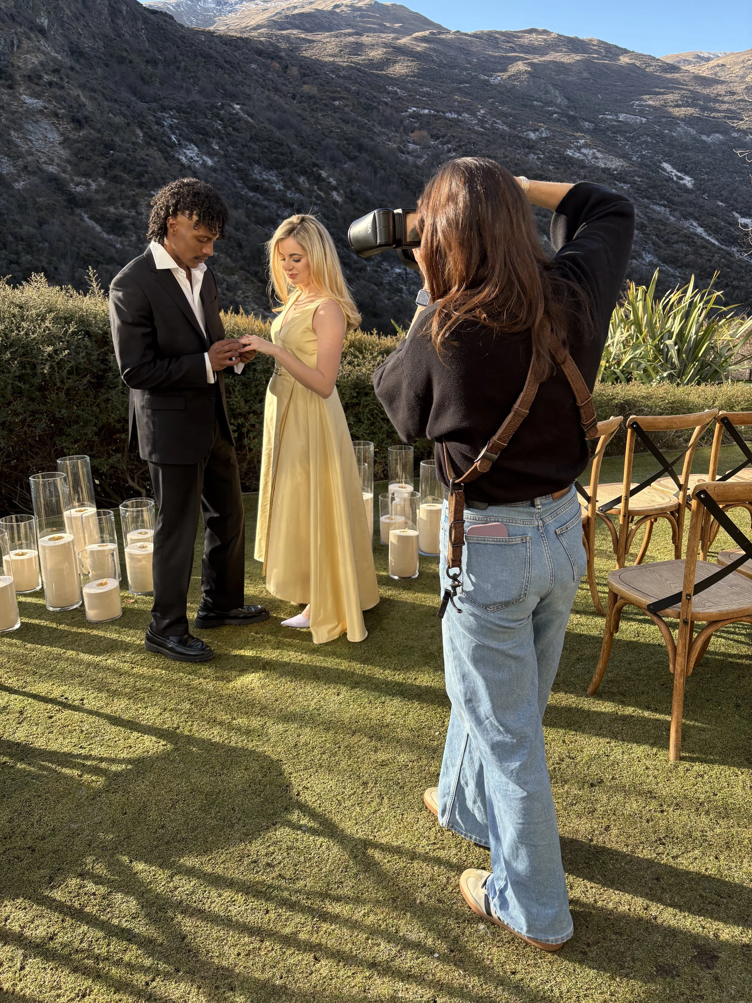 A woman is taking a photograph of a couple exchanging rings during an outdoor wedding ceremony on a grassy area with mountains in the background. Candles are arranged on the ground and chairs are set up nearby.