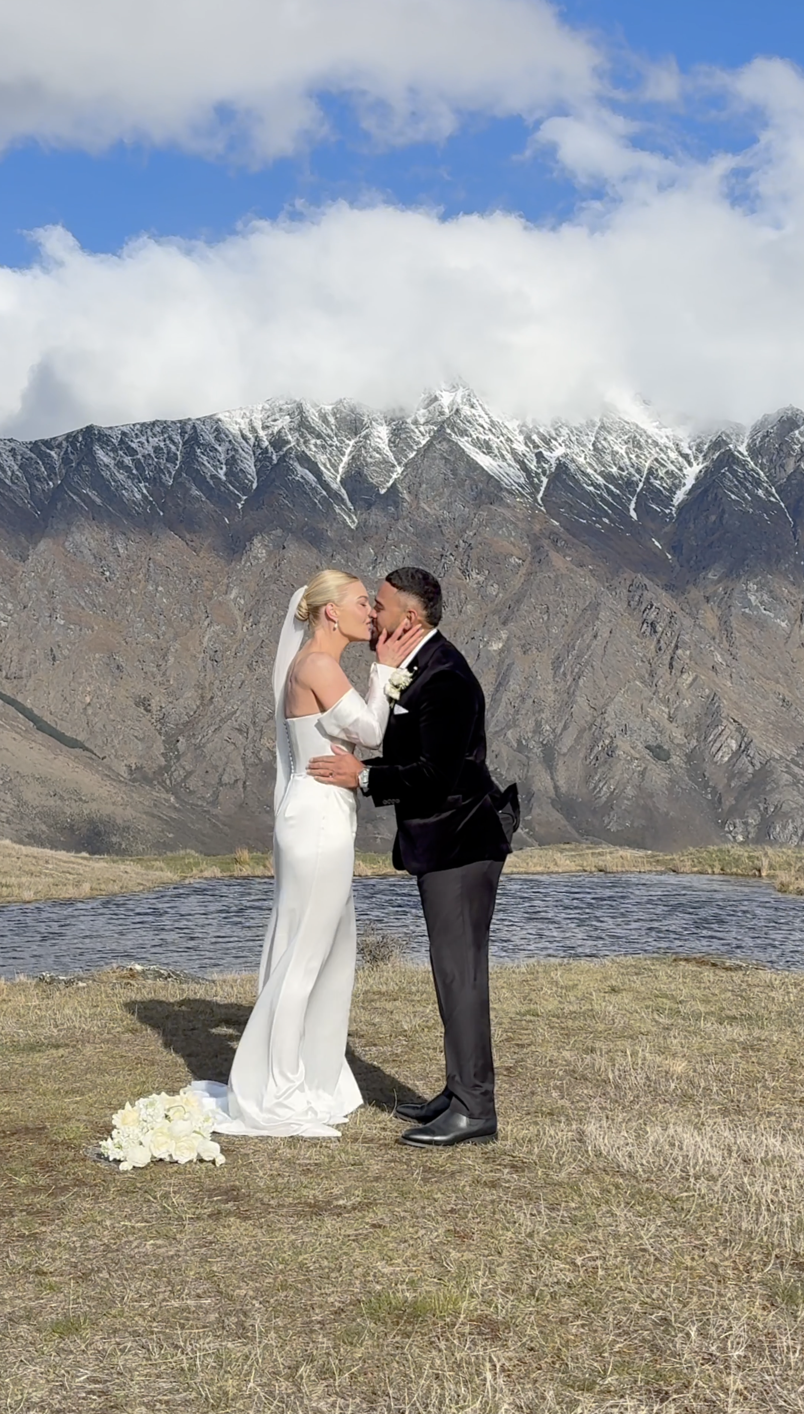 A bride and groom sharing a kiss outdoors with mountains and a lake in the background. Auckland content creators